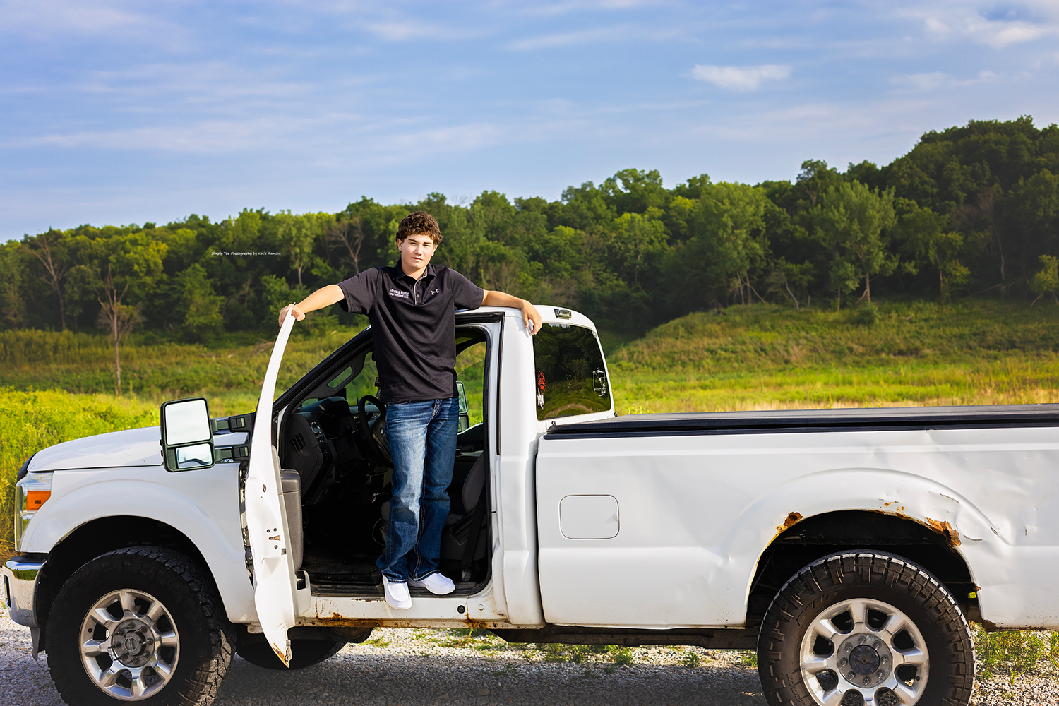 Young man standing in open door of white pickup truck parked on rural road with green fields behind.