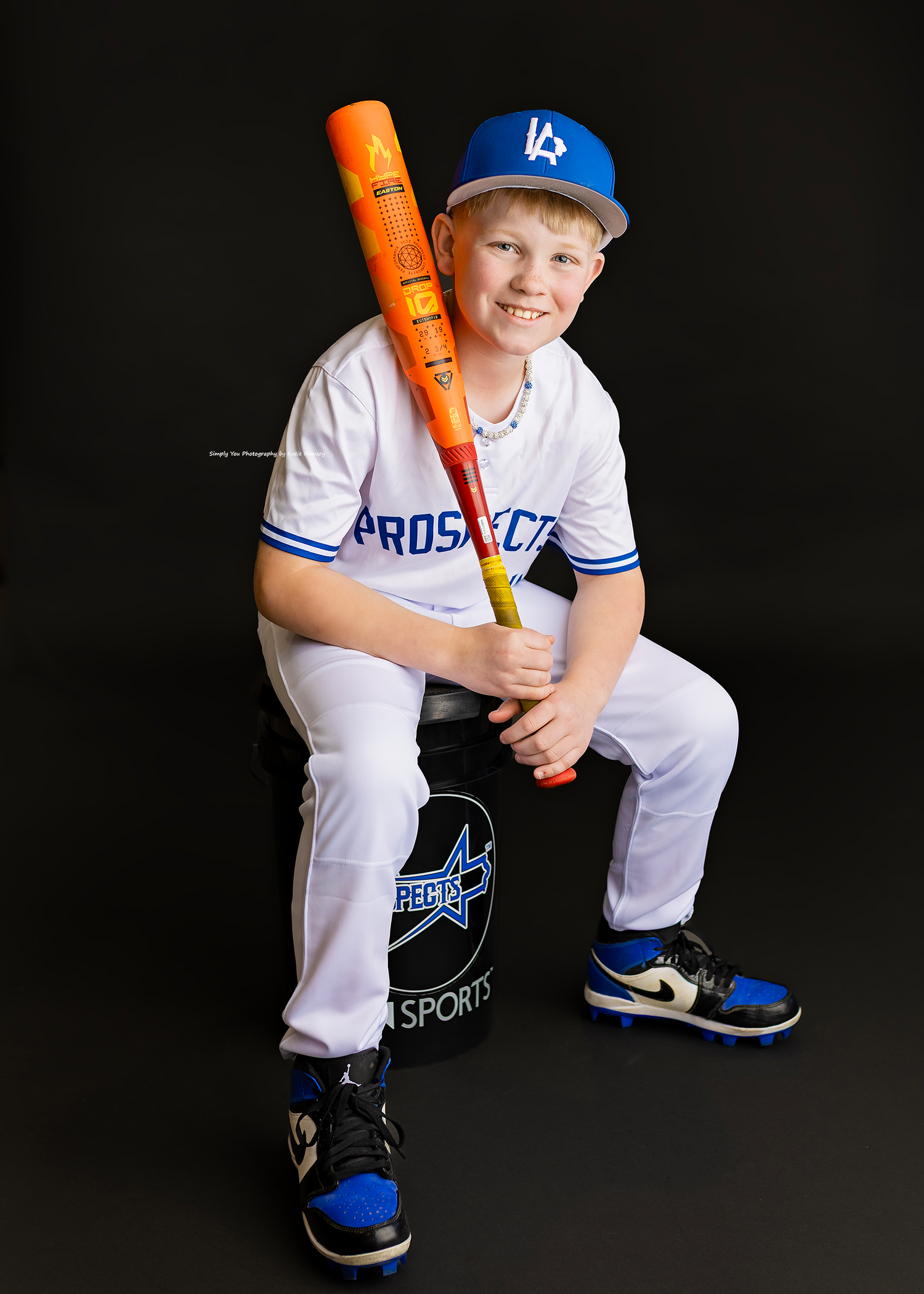 Young baseball player in Prospects uniform and LA Dodgers cap poses with orange bat on sports bag.