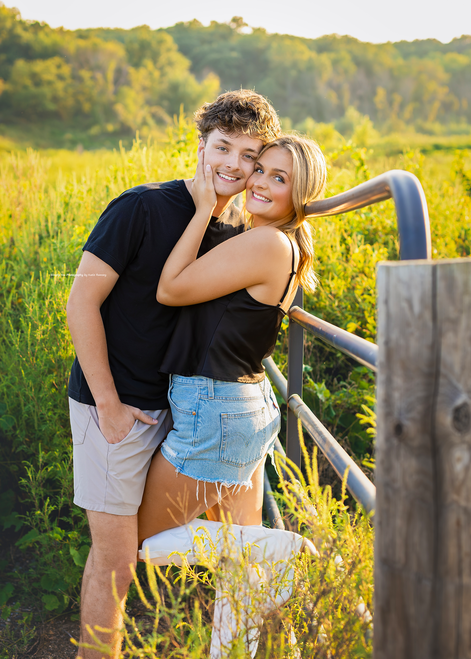 A young couple smiles together by a rustic fence in a sunny green field, wearing casual black tops and shorts.