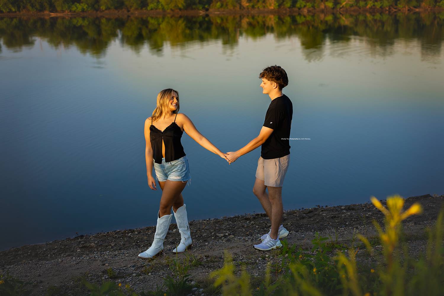 A young couple holds hands by a calm lake at sunset, she wears boots and denim shorts, he wears black shirt.