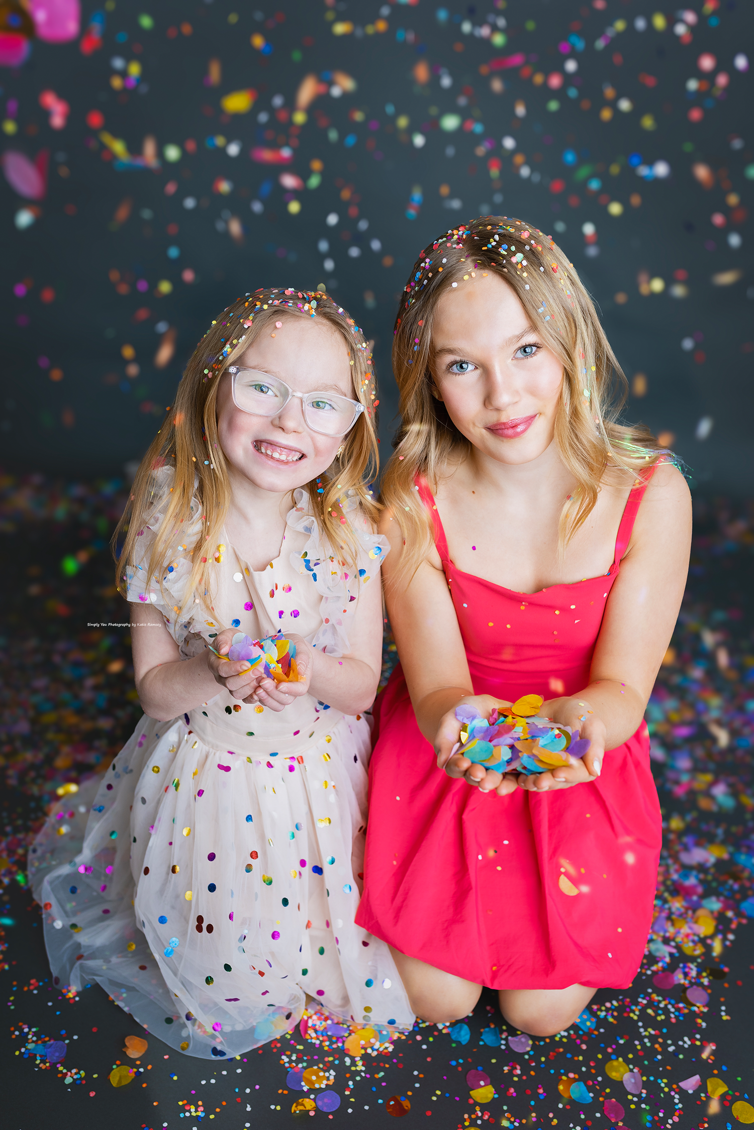 Two sisters kneeling together holding confetti, smiling in a colorful confetti-filled photography studio.