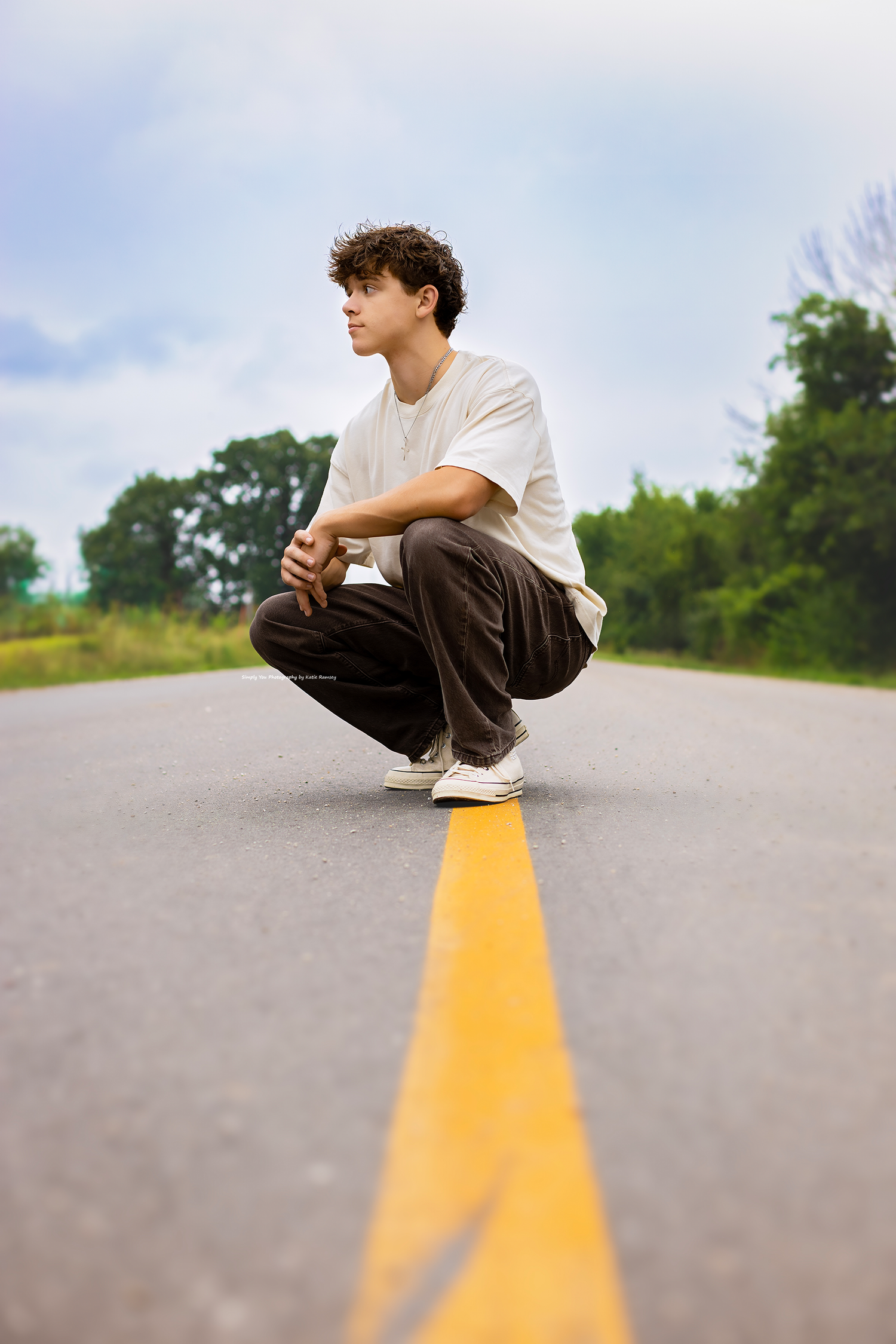 Young man with curly hair crouching on a road centerline, wearing white tee and brown pants, trees in background.