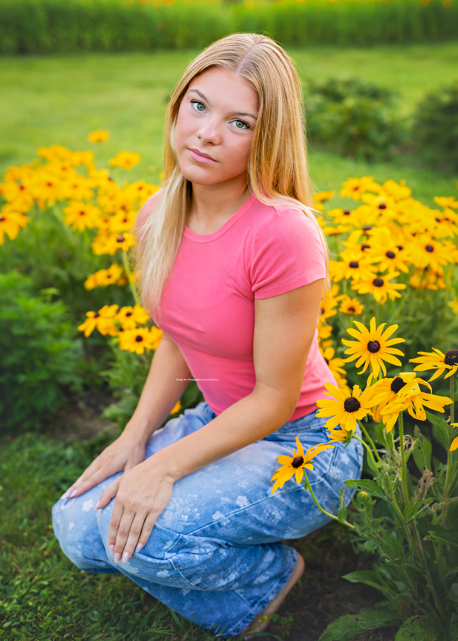 Young blonde woman in pink shirt and jeans crouching among bright yellow black-eyed Susan flowers in a lush green garden.