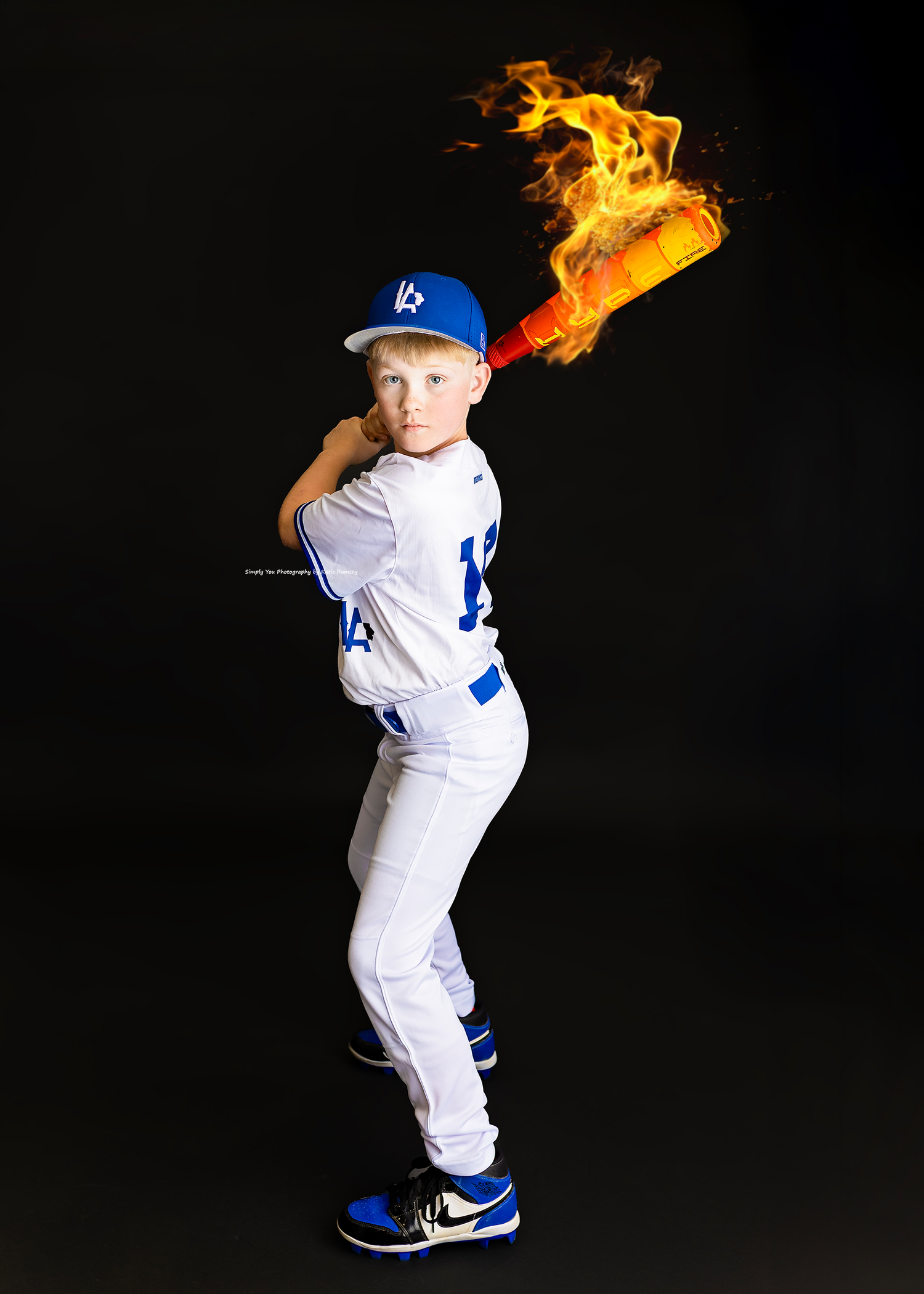 Young baseball player in Dodgers uniform poses with a flaming bat against a black background.