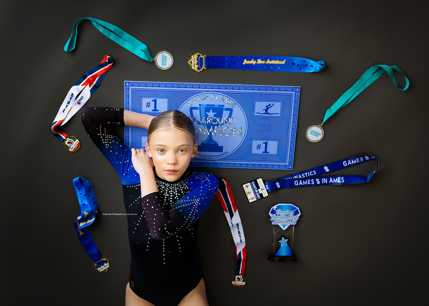 Young gymnast poses with blue leotard surrounded by medals, trophies, and competition ribbons on dark background.