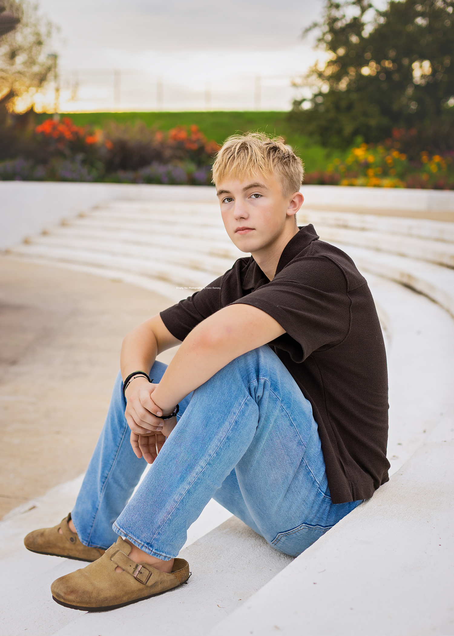A young blonde teen sits casually on white steps wearing jeans, a black shirt, and tan clogs in an outdoor setting.