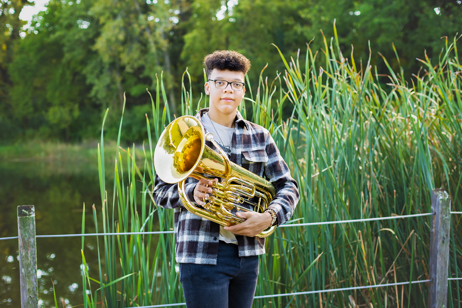 Young musician holding a French horn outdoors near a pond with tall green reeds and trees in the background.