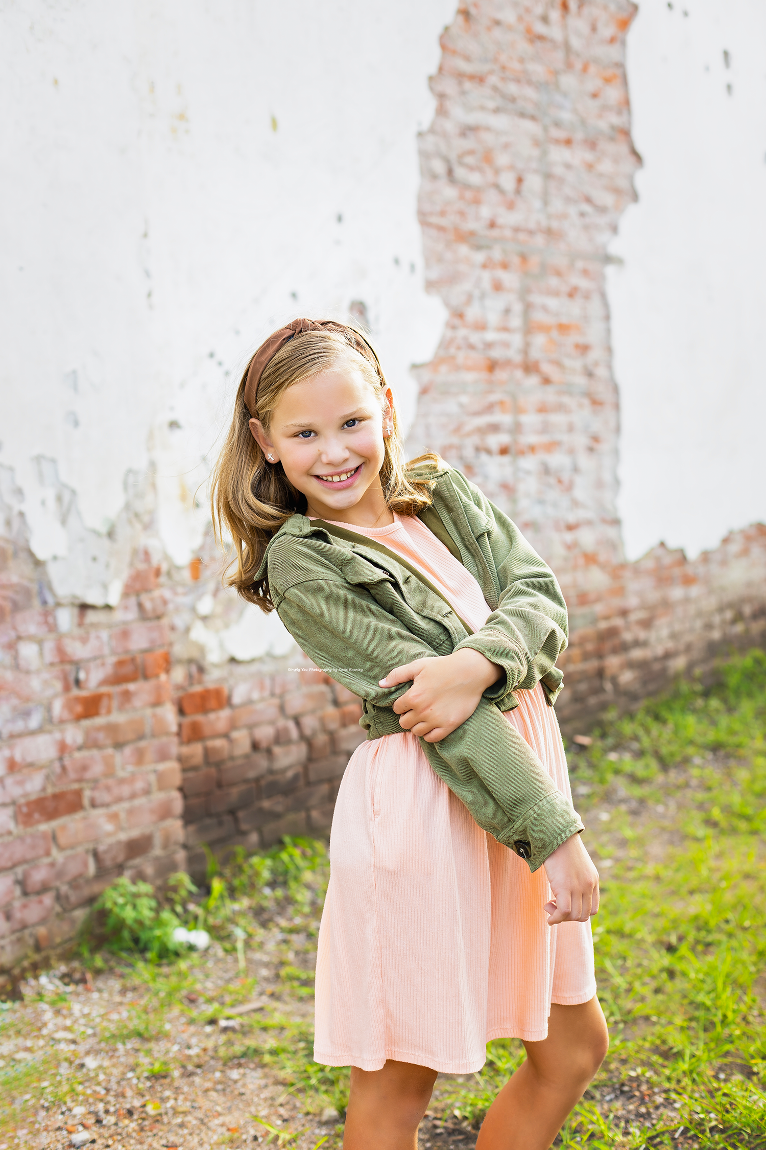 A smiling young girl in a pink dress and green jacket poses outdoors near a rustic brick wall.