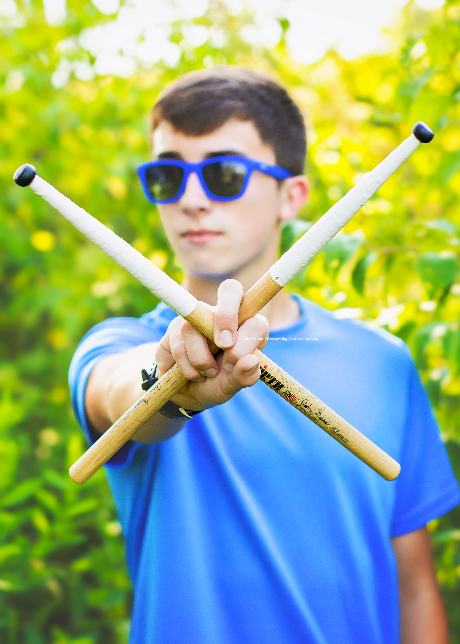 Teen boy in blue shirt and sunglasses holding crossed drumsticks outdoors.