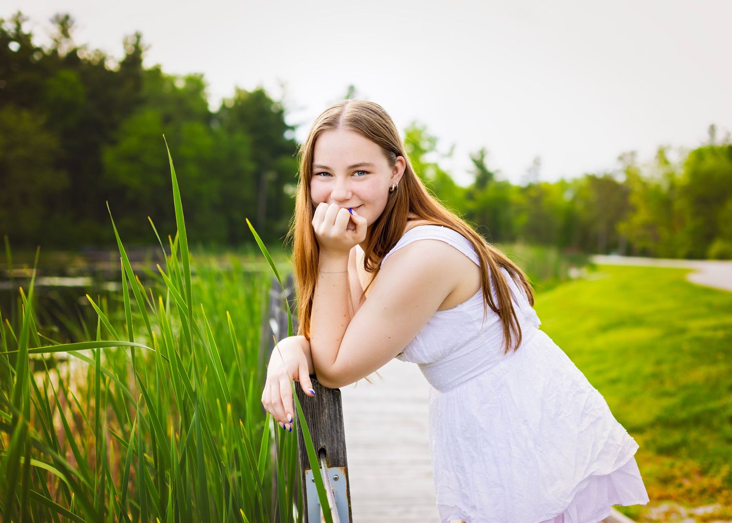 Smiling young woman in white dress leaning on fence near tall grass in a lush green park.
