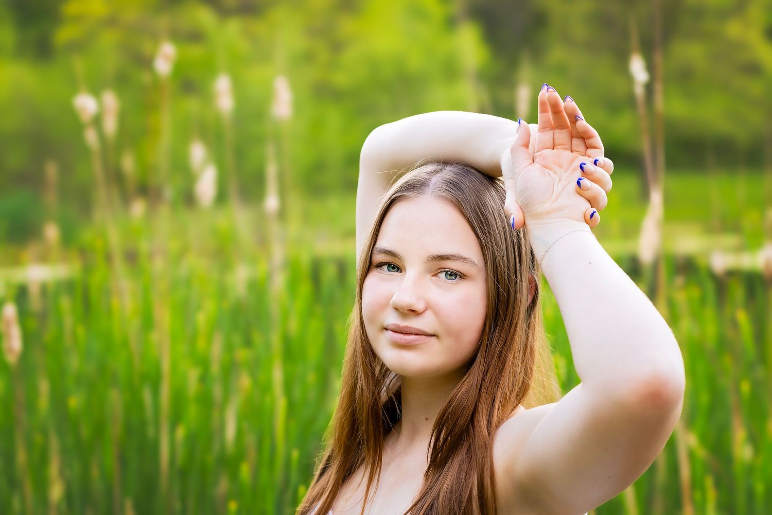 Young woman with long brown hair poses outdoors near lush green reeds, hand raised gracefully above her head.