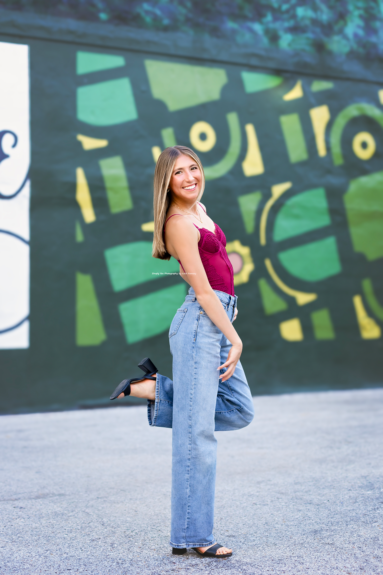 Smiling young woman in burgundy top and wide-leg jeans posing in front of green geometric mural.