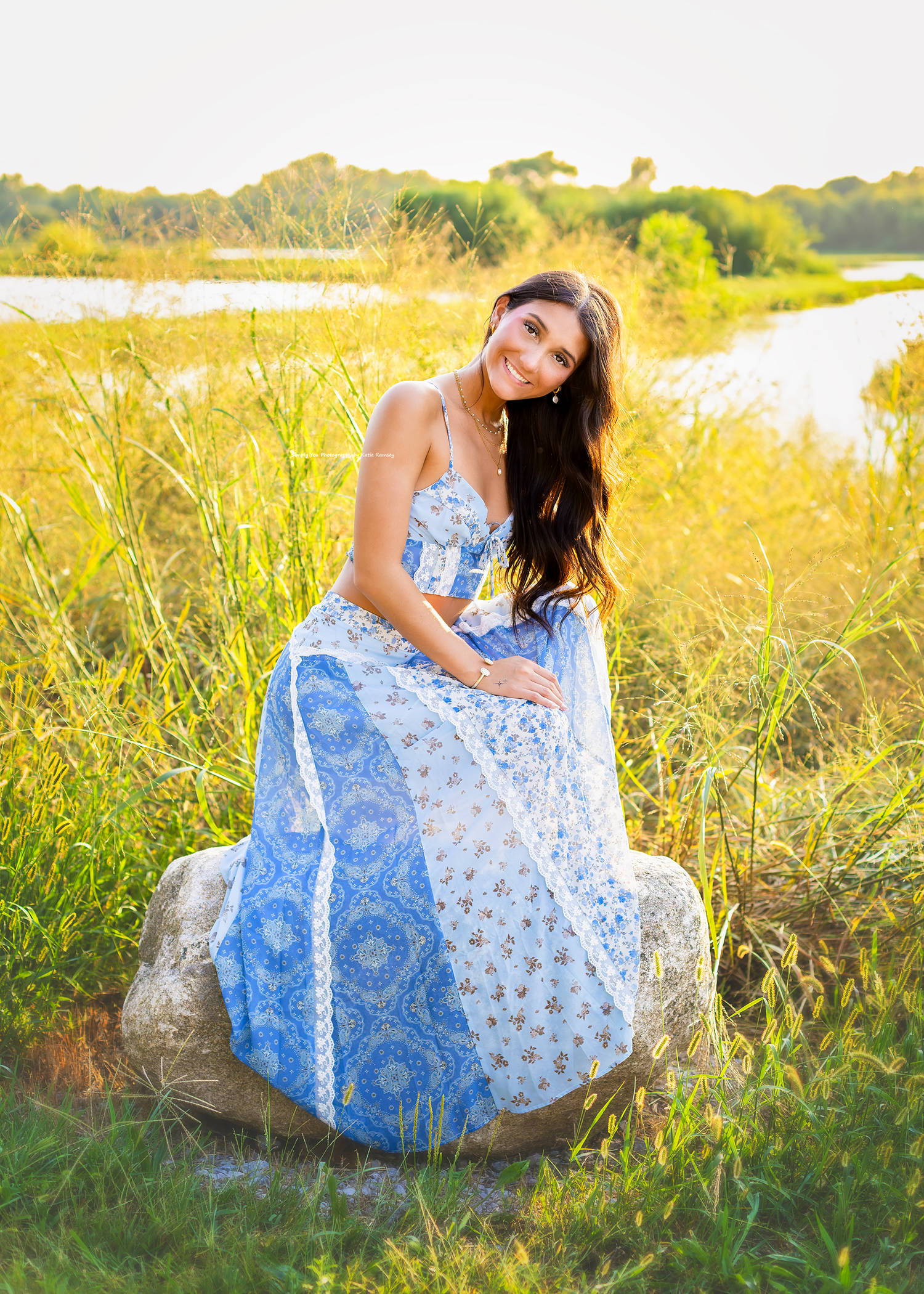 Young woman in blue floral maxi dress sitting on rock surrounded by golden tall grass near water.