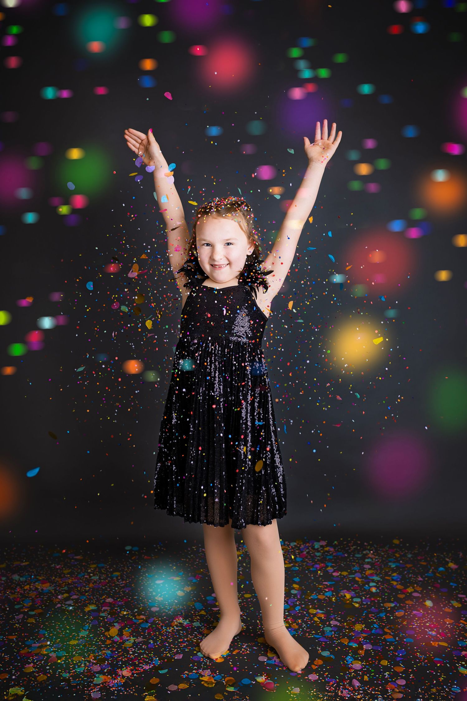 Happy girl in black velvet dress jumping with arms raised amid falling confetti.