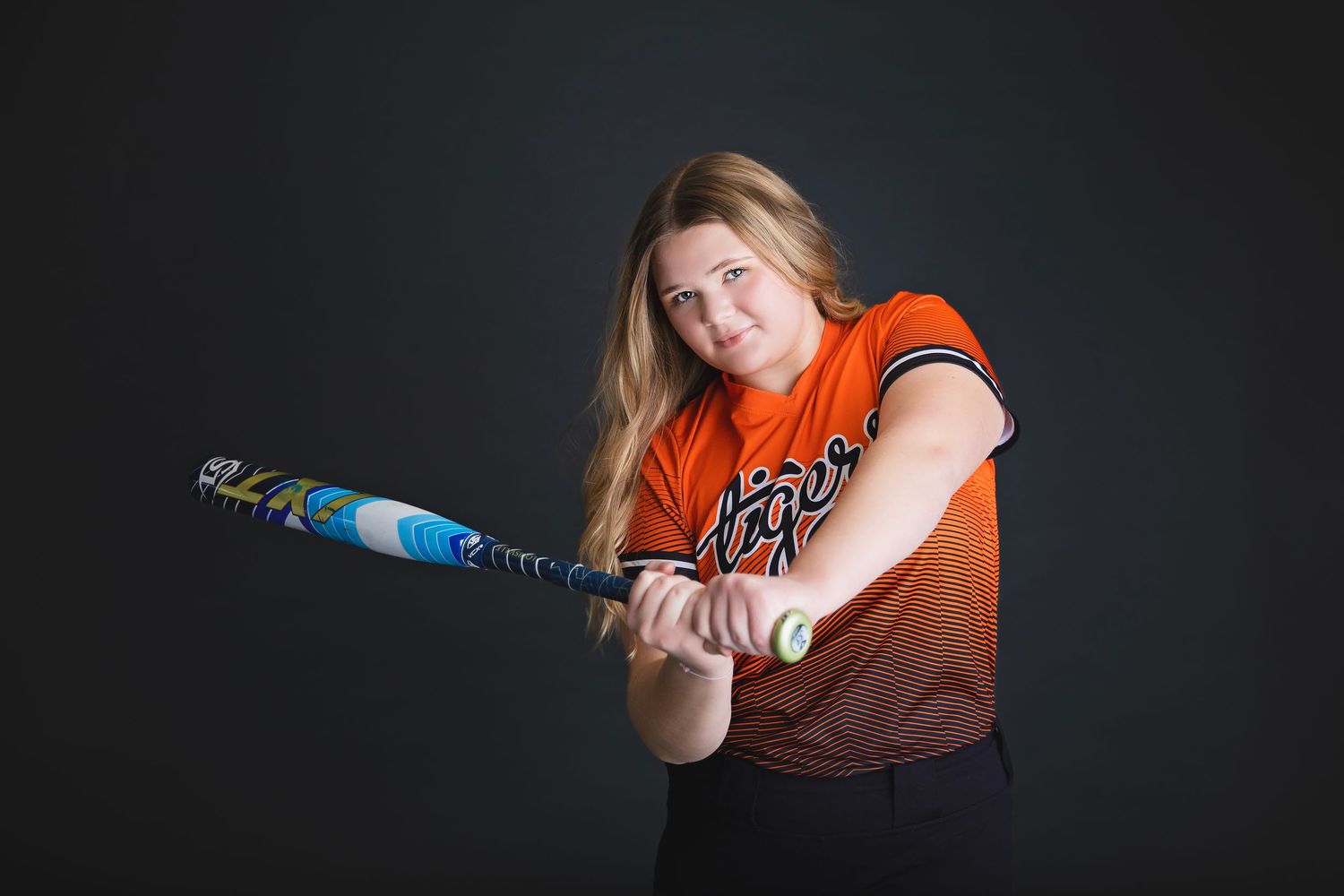 Teen softball player in orange Tigers jersey swings bat in action pose against dark background.