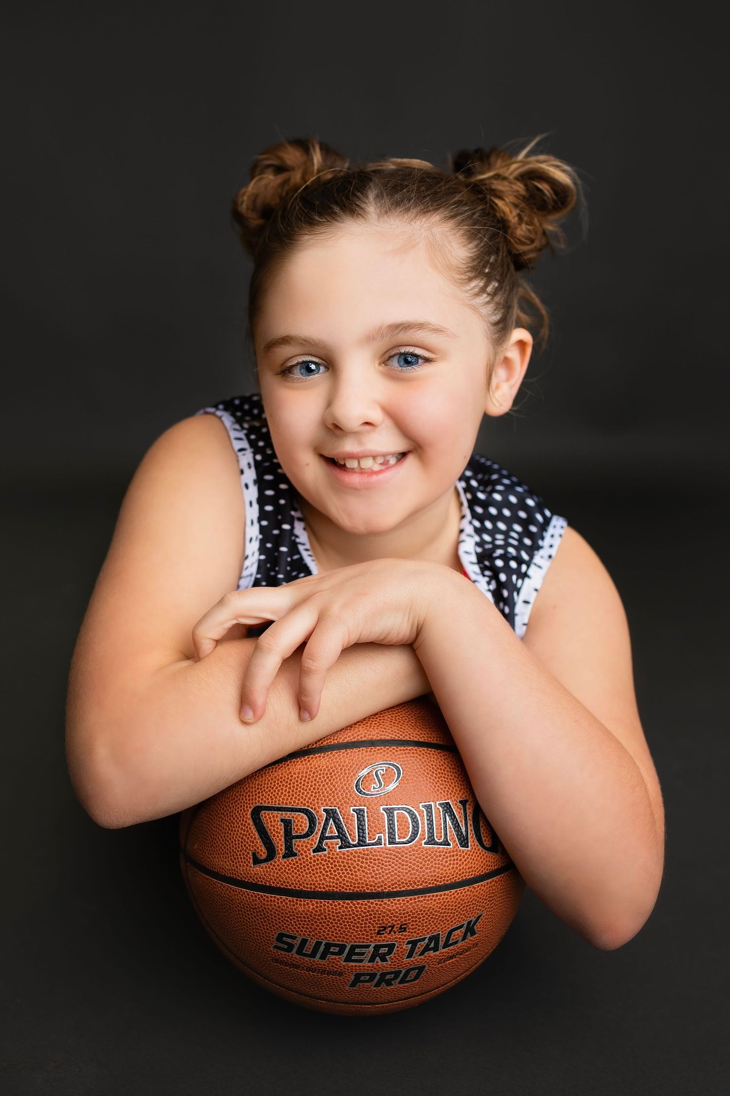 Young girl smiling with blue eyes leaning on a Spalding Super Tack Pro basketball against dark background.