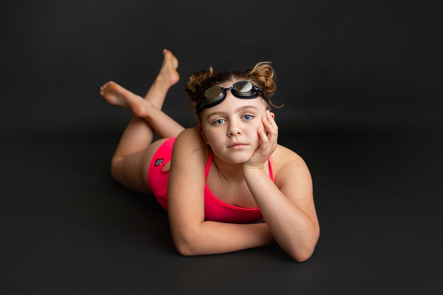 Young girl in pink swimsuit with goggles lying on floor against dark background looking at camera.