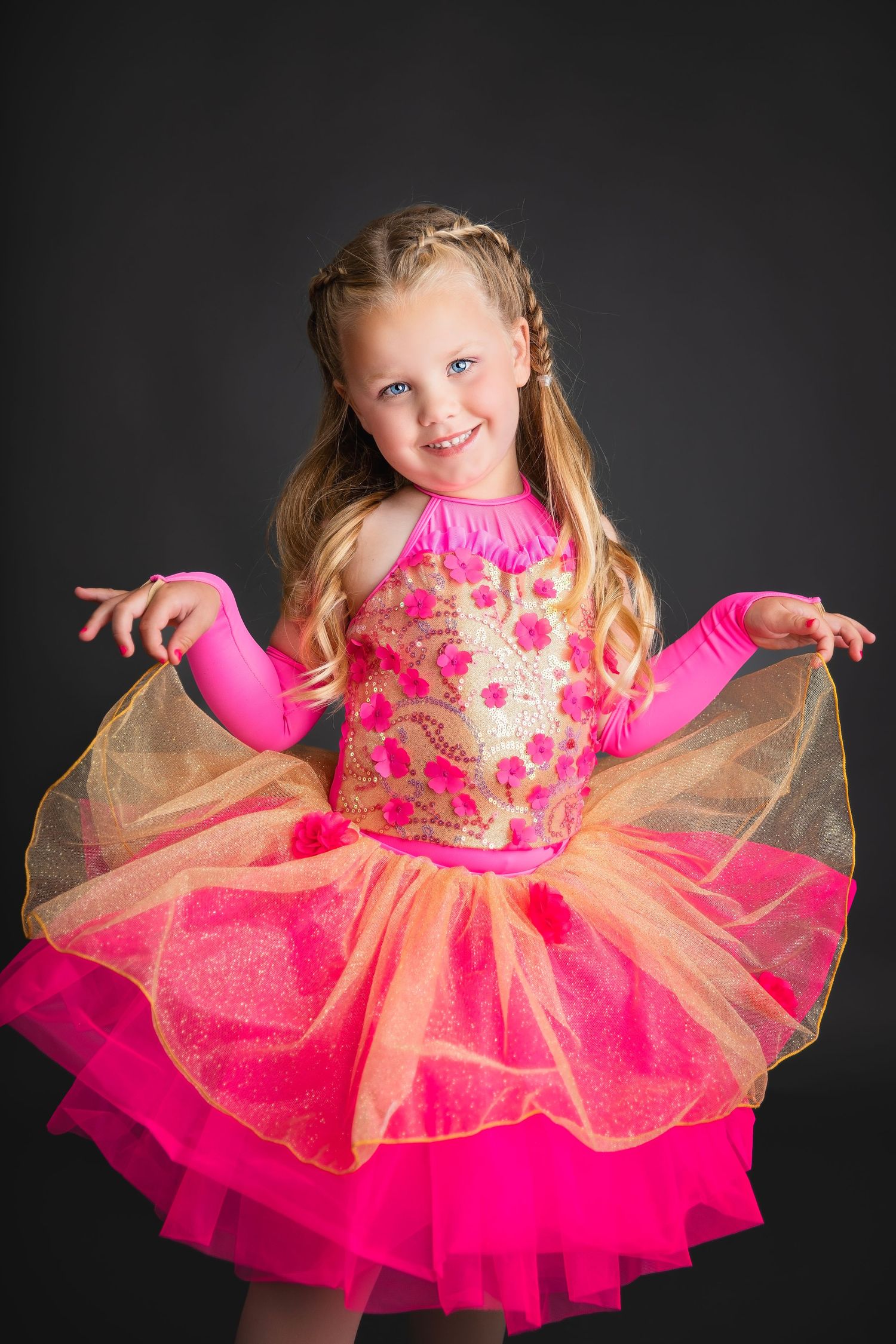 Young girl smiling in pink and orange sequined tutu dance costume holding out skirt in studio.
