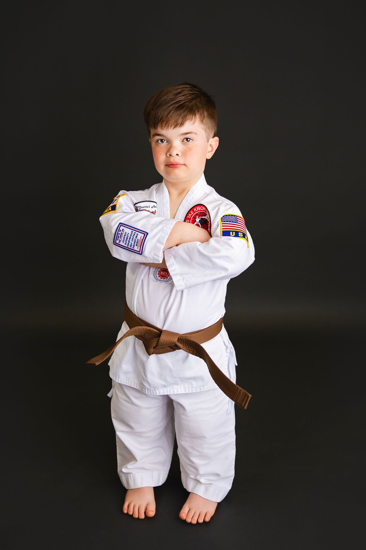 Young boy in white karate gi with brown belt stands with arms crossed in studio.
