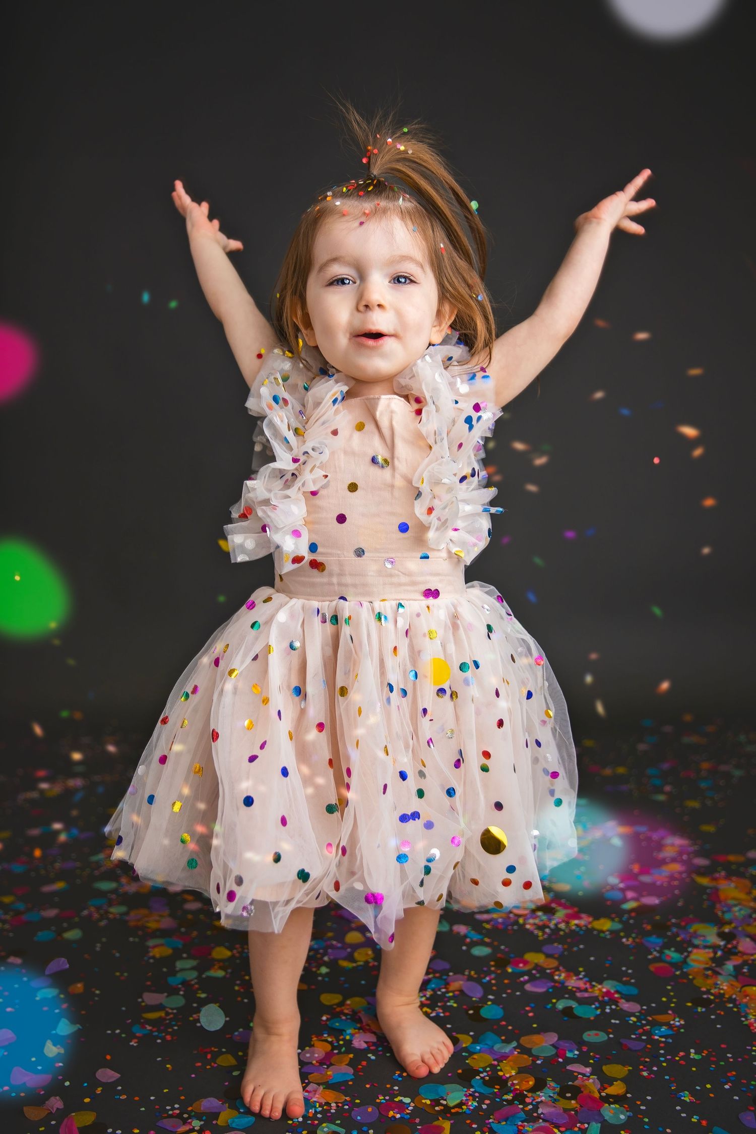 Excited toddler girl in polka dot tulle dress with arms raised, surrounded by falling confetti.