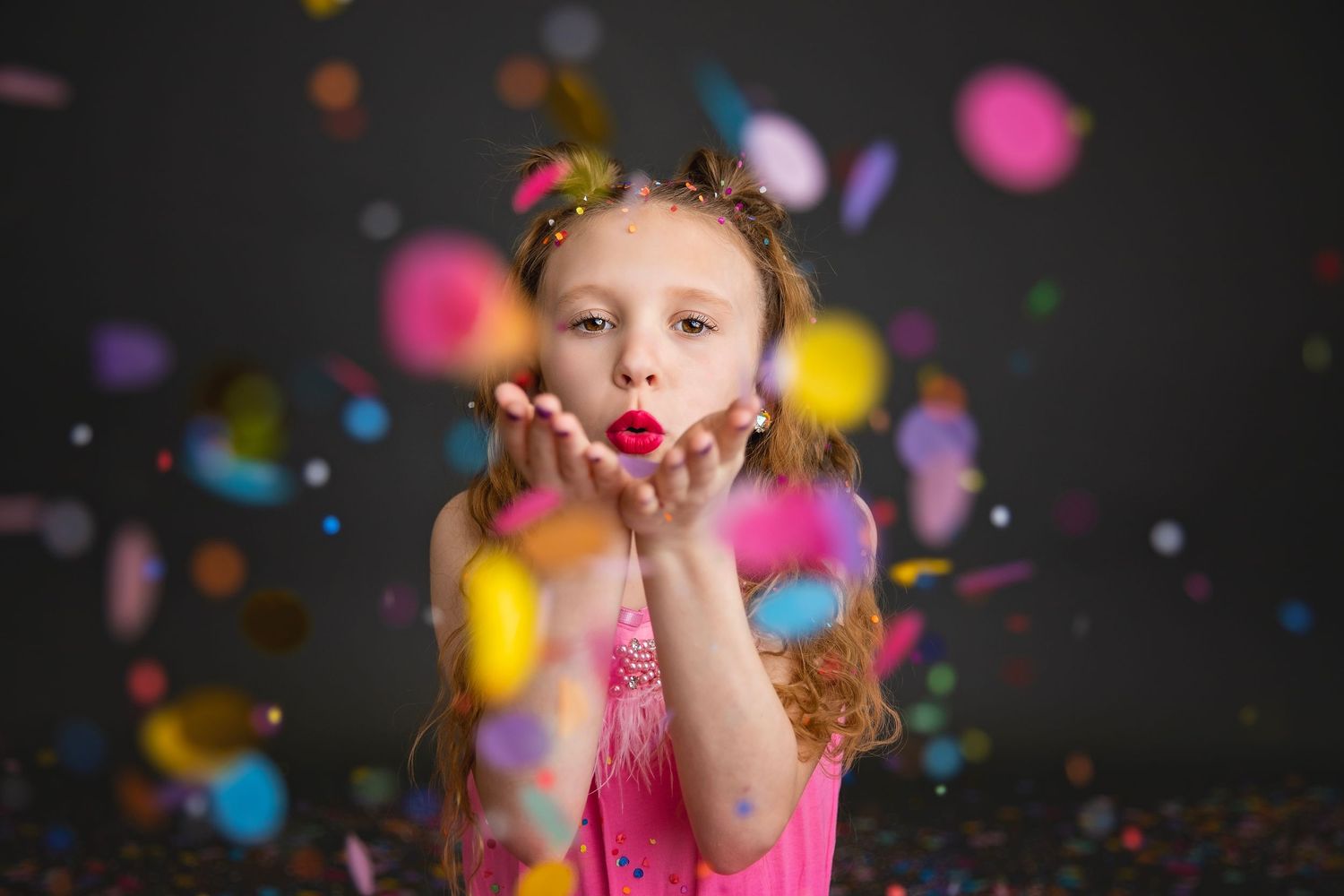 Young girl in pink dress blowing colorful confetti from her hands.
