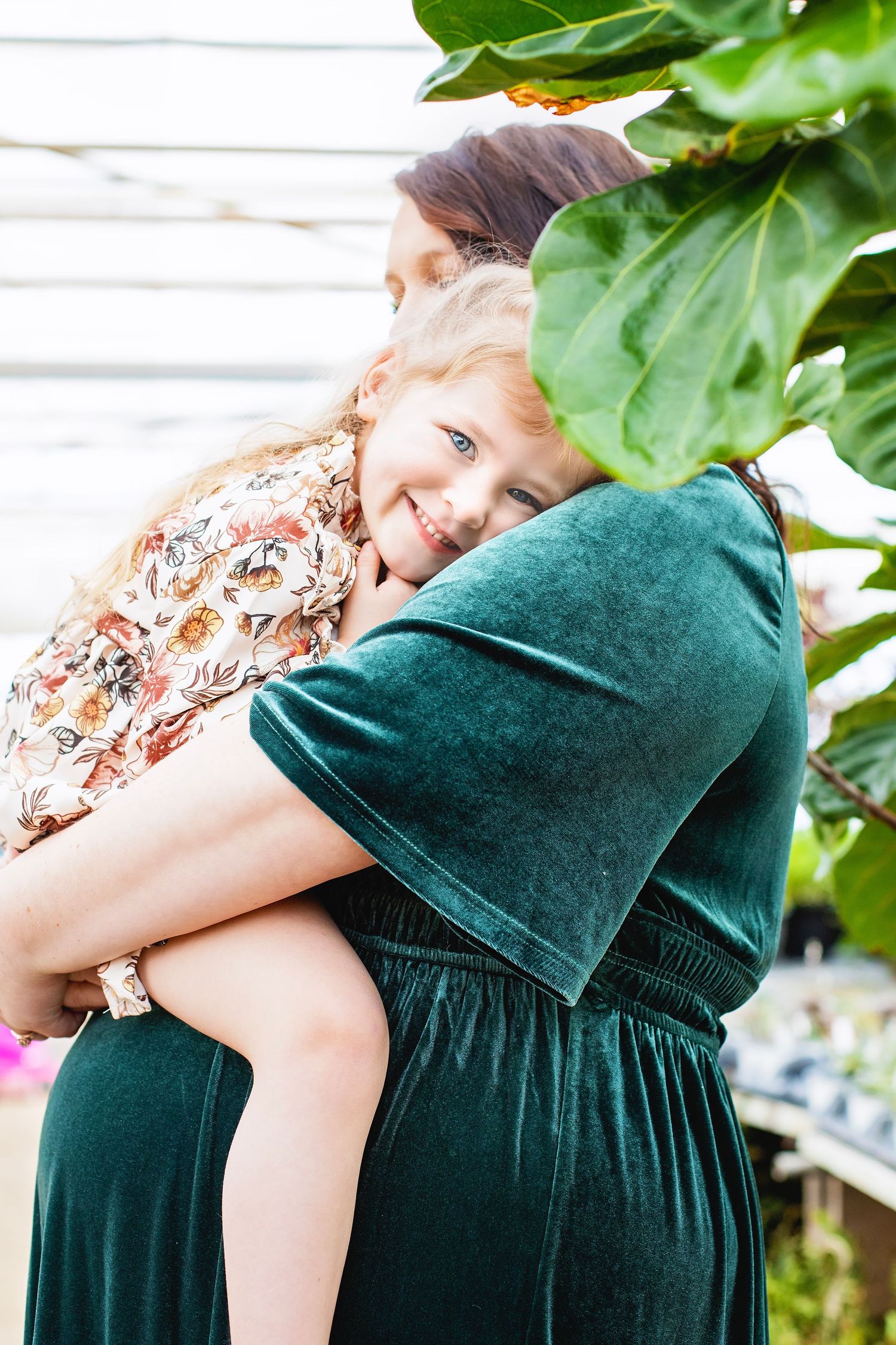 A smiling young girl with curly blonde hair is held by a woman in a green velvet dress near tropical plants.