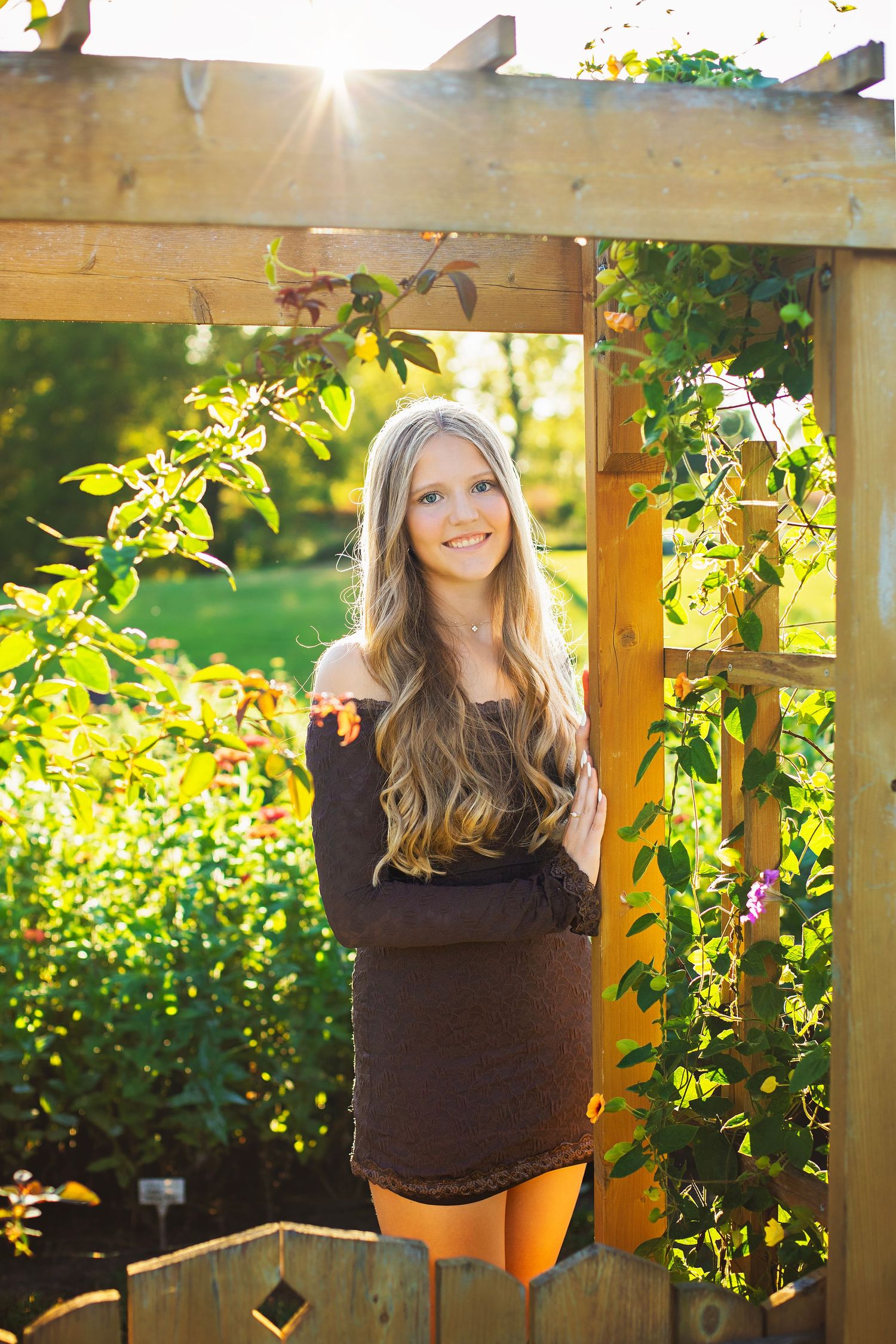 Smiling blonde teen in dark off-shoulder dress stands under wooden garden pergola in golden sunlight.