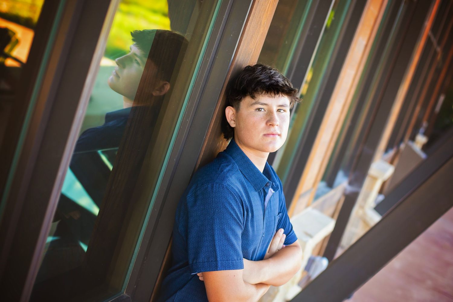 Teen boy with arms crossed leaning against glass panels reflecting colorful columns outdoors.