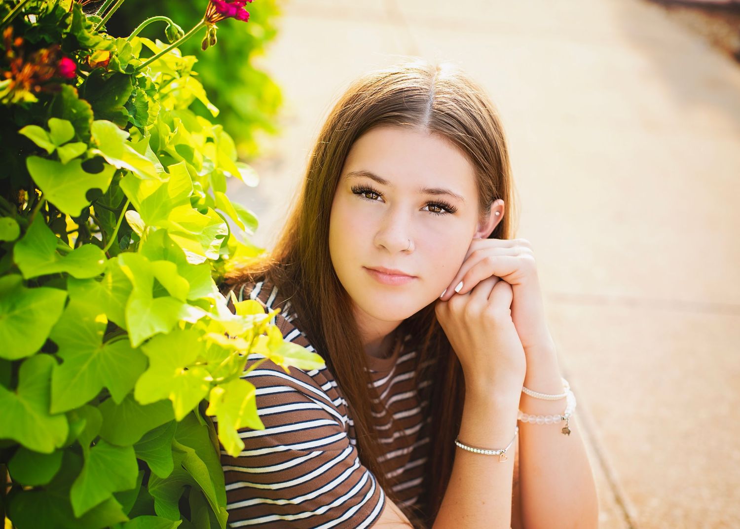 Pretty teen girl with long brown hair resting chin on hand near colorful garden flowers.