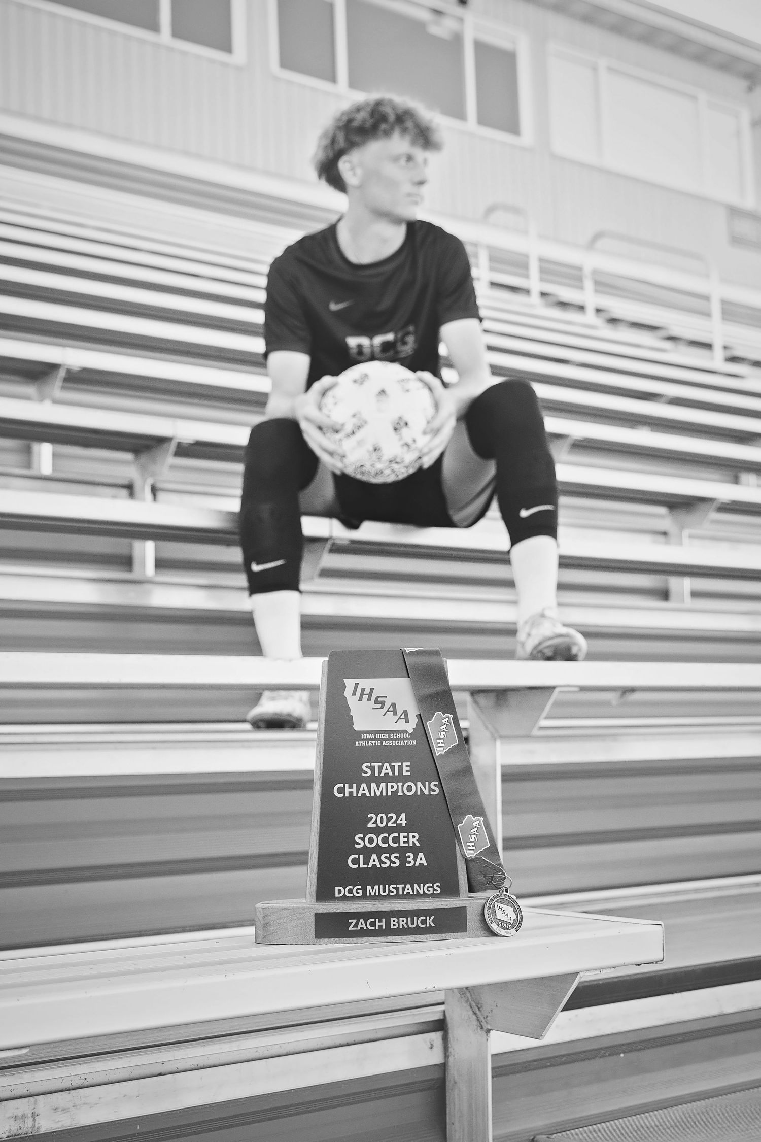 Black and white photo of soccer player with 2024 IHSAA State Champions Class 3A trophy in foreground.