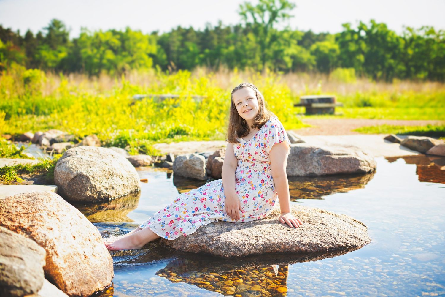 Young girl in floral dress sitting on rocks near peaceful stream in sunny outdoor setting.