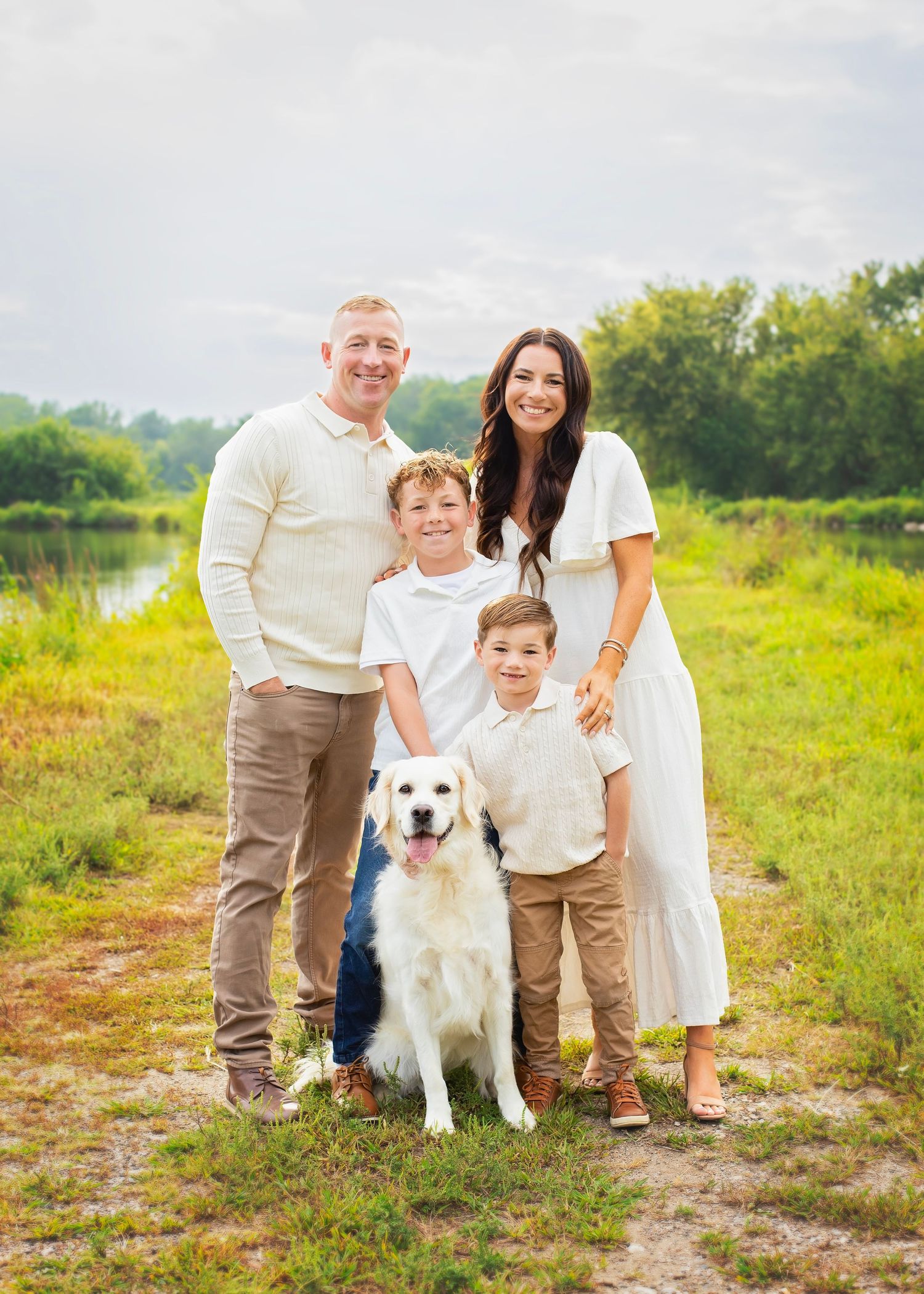 Family of four with a golden retriever dog posing outdoors in a green meadow near a pond.