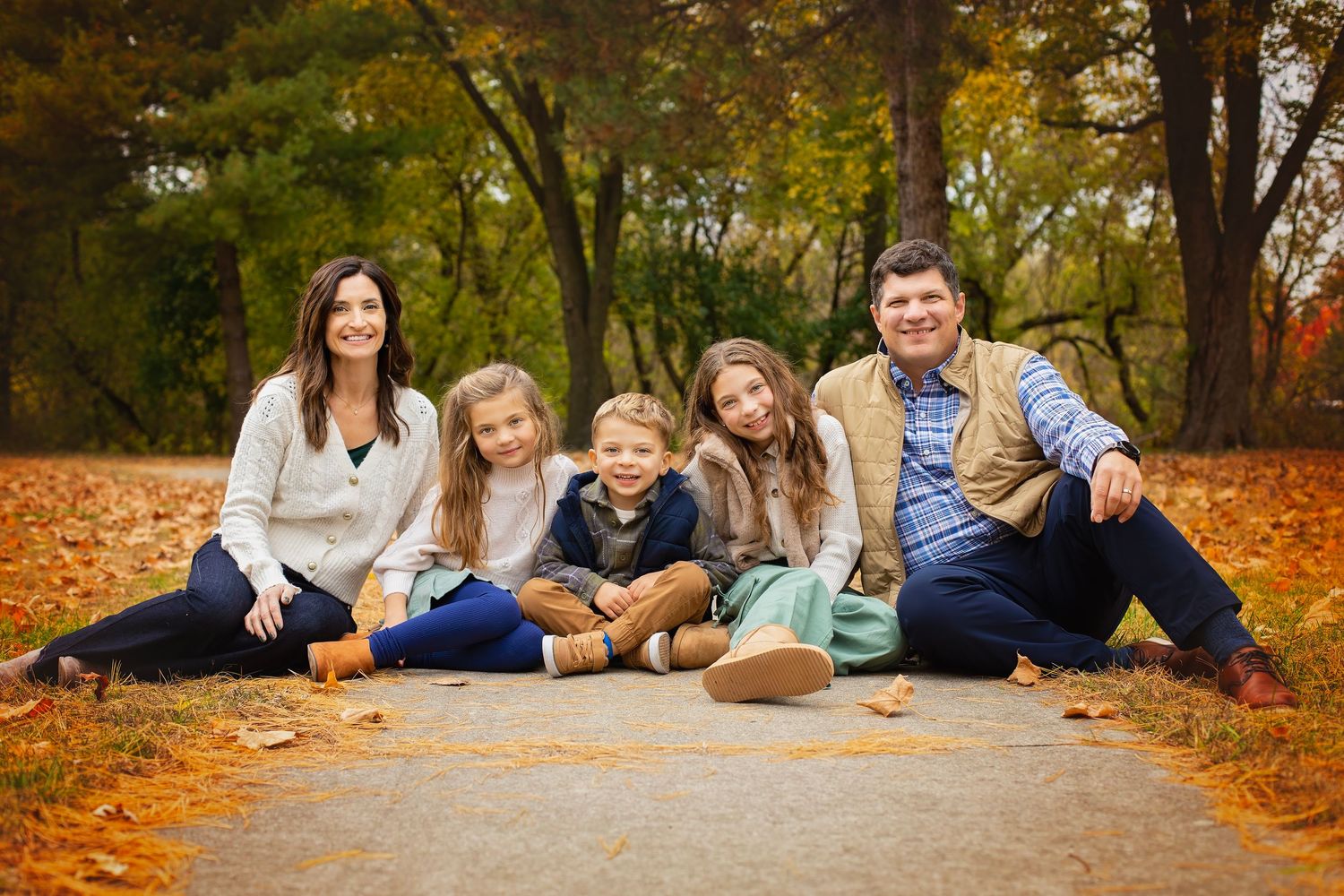 A happy family of five sits together on a leaf-covered path surrounded by colorful autumn trees.