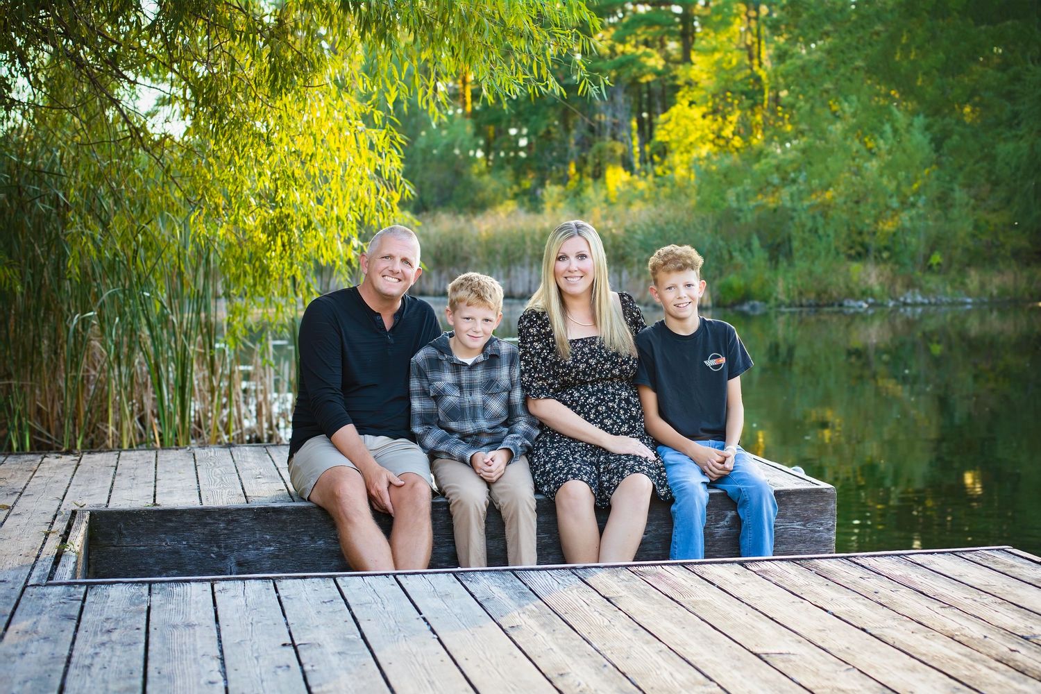 A happy family of four sits together on a wooden dock surrounded by lush green trees by a pond.