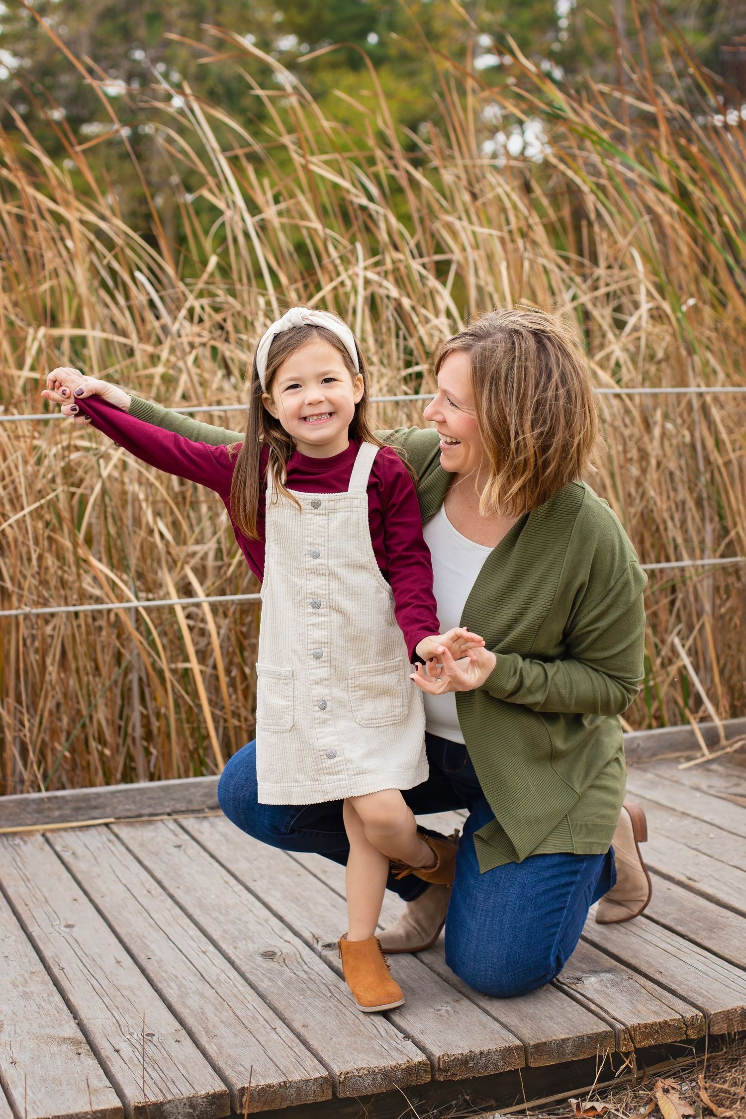 Mother kneeling beside smiling young daughter on a wooden deck with tall grass background.