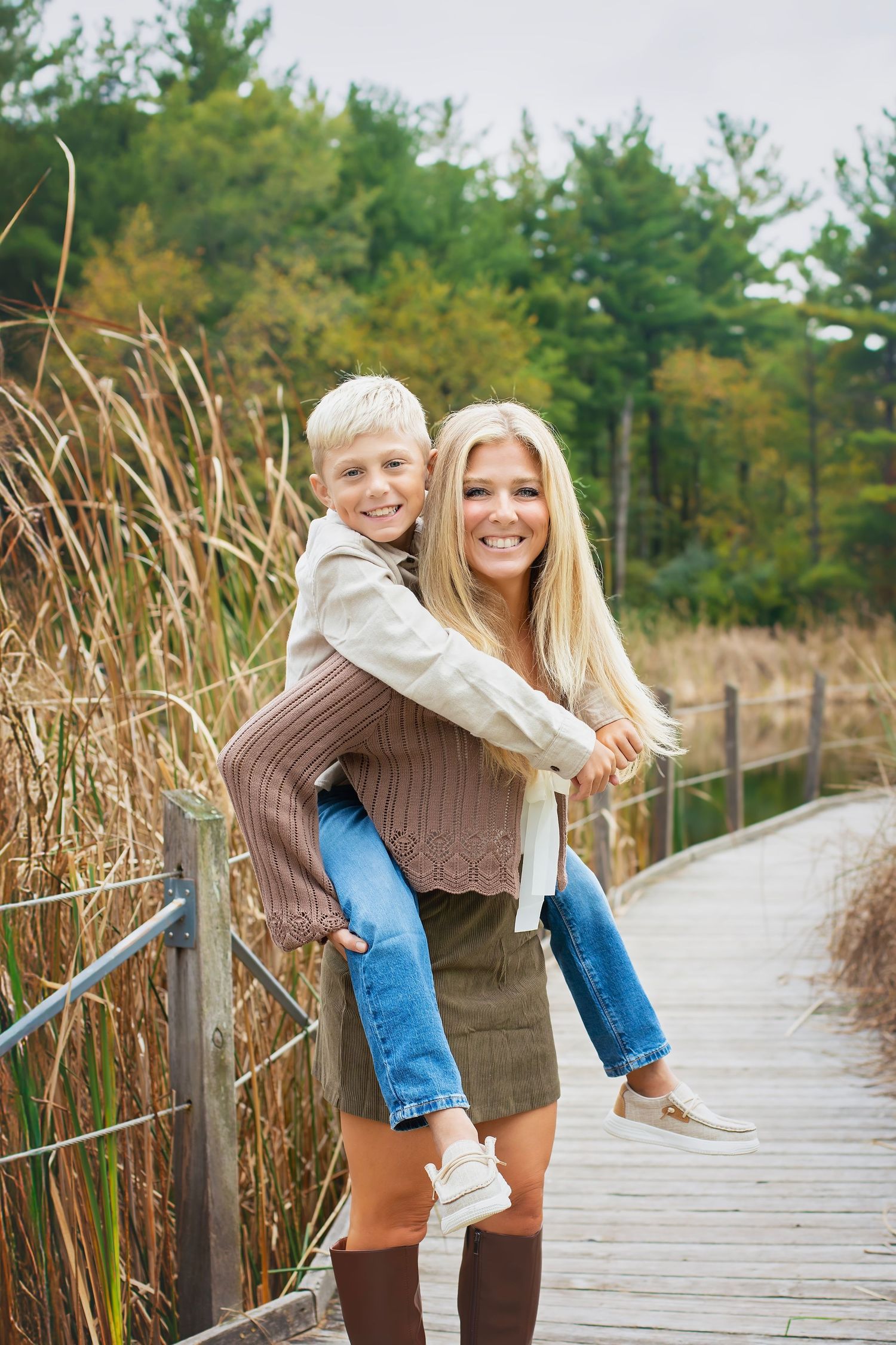 A blonde woman gives her young son a piggyback ride on a wooden boardwalk surrounded by tall autumn grasses.