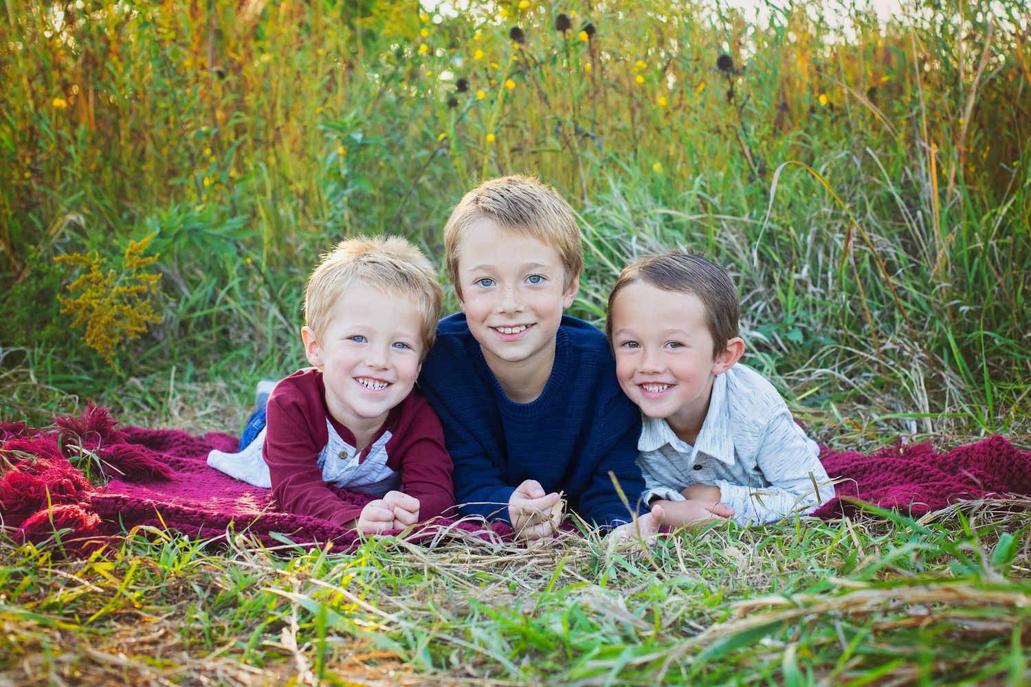 Three smiling brothers lying on a red blanket in a lush green meadow.