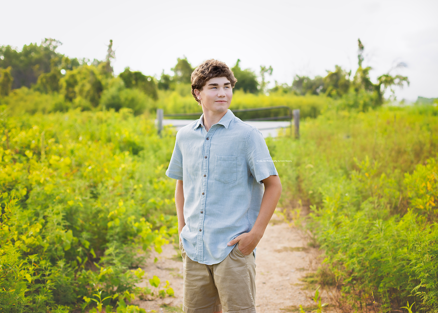 A teenage boy in a light blue shirt and khaki pants stands on a dirt path surrounded by lush green foliage.