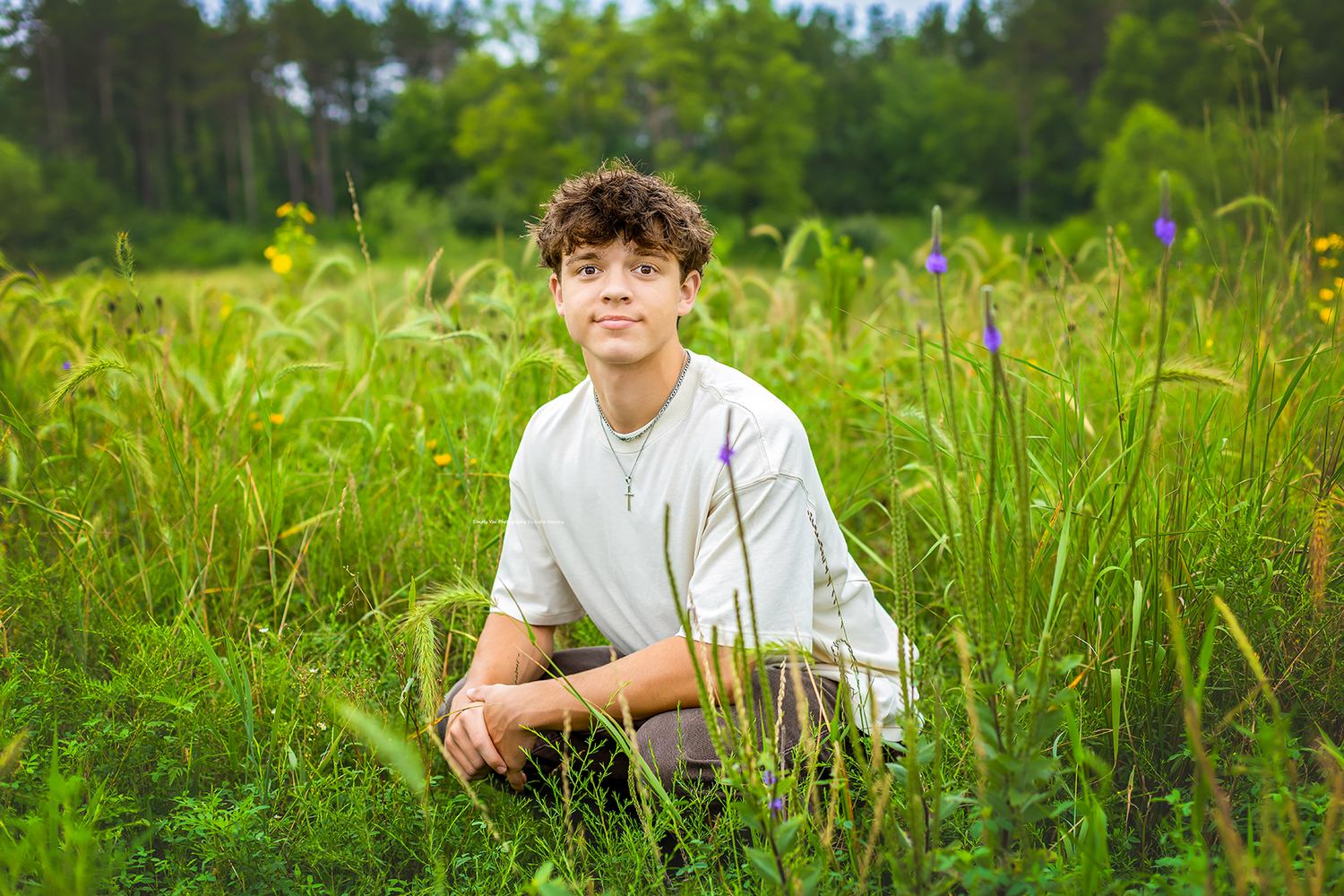 Teen boy with curly hair sits in lush green meadow with wildflowers during outdoor senior portrait session.