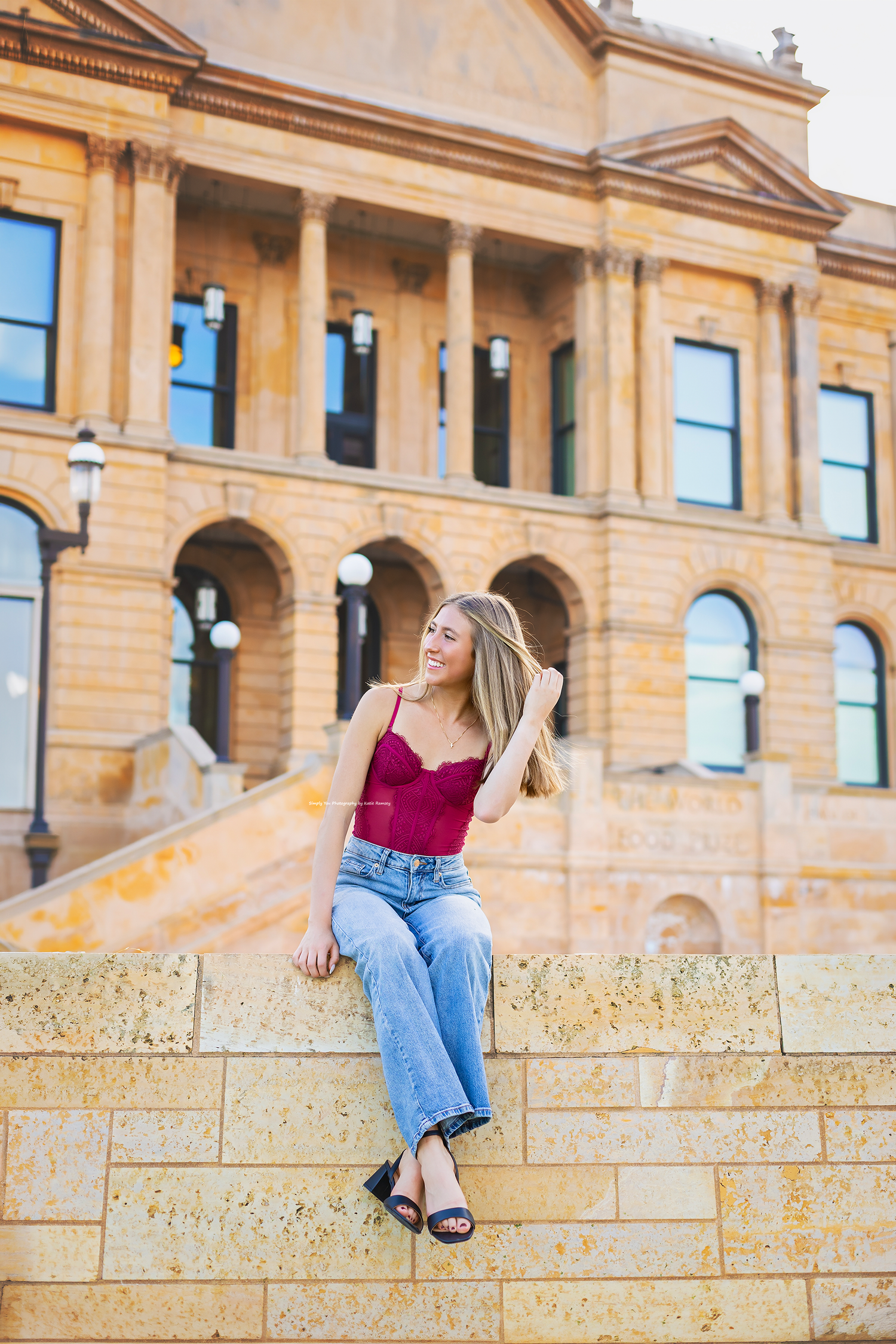 Blonde woman in red top and jeans sits on stone wall outside classic sandstone building.