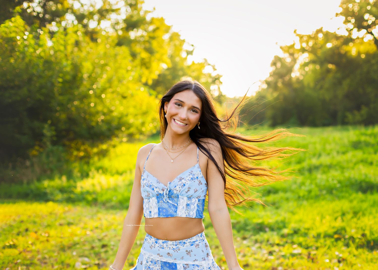 Young woman in blue floral crop top smiling in a sunny green meadow with hair blowing in the wind.