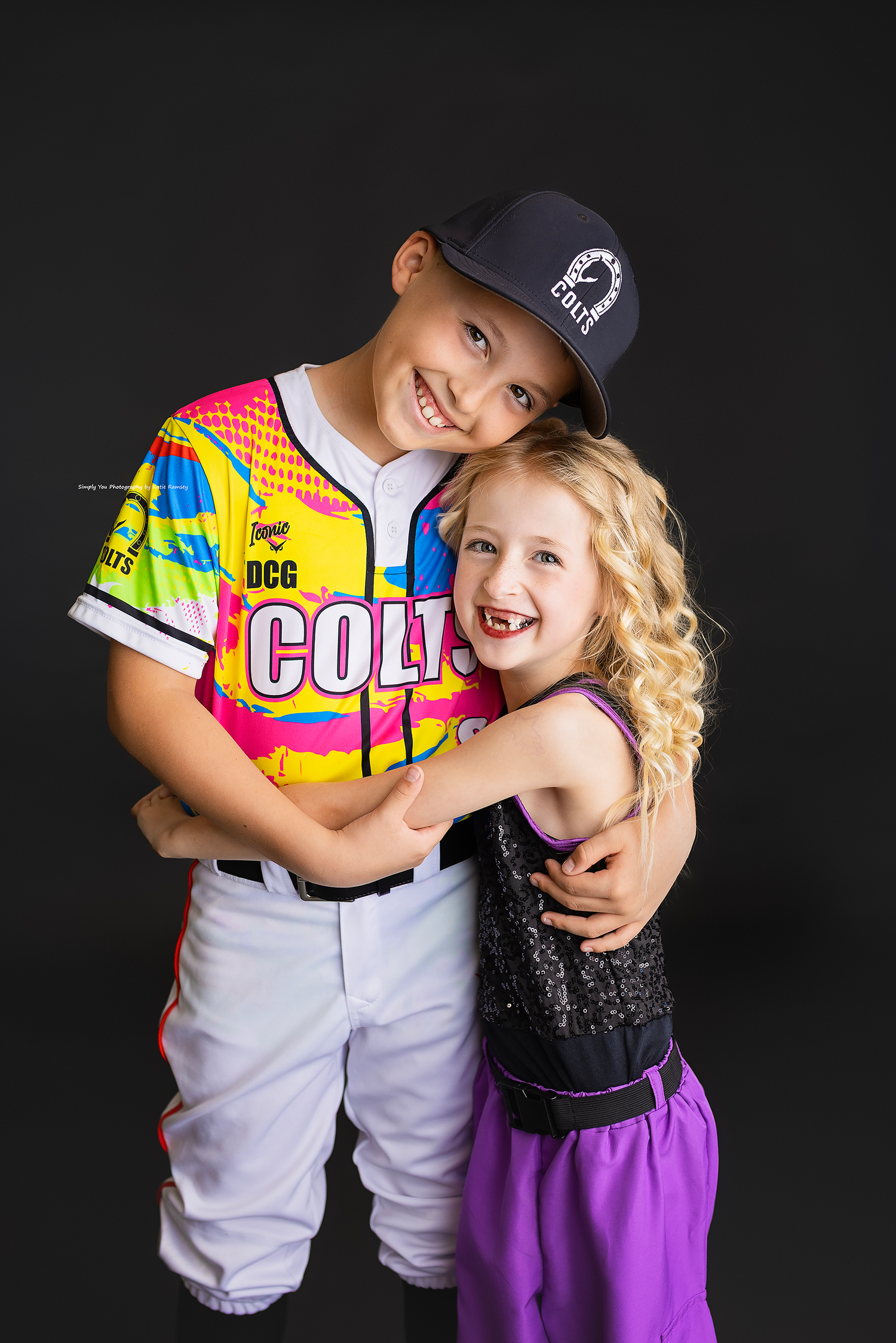 Two children hug and smile, boy in colorful Colts baseball jersey and girl in purple dance outfit.