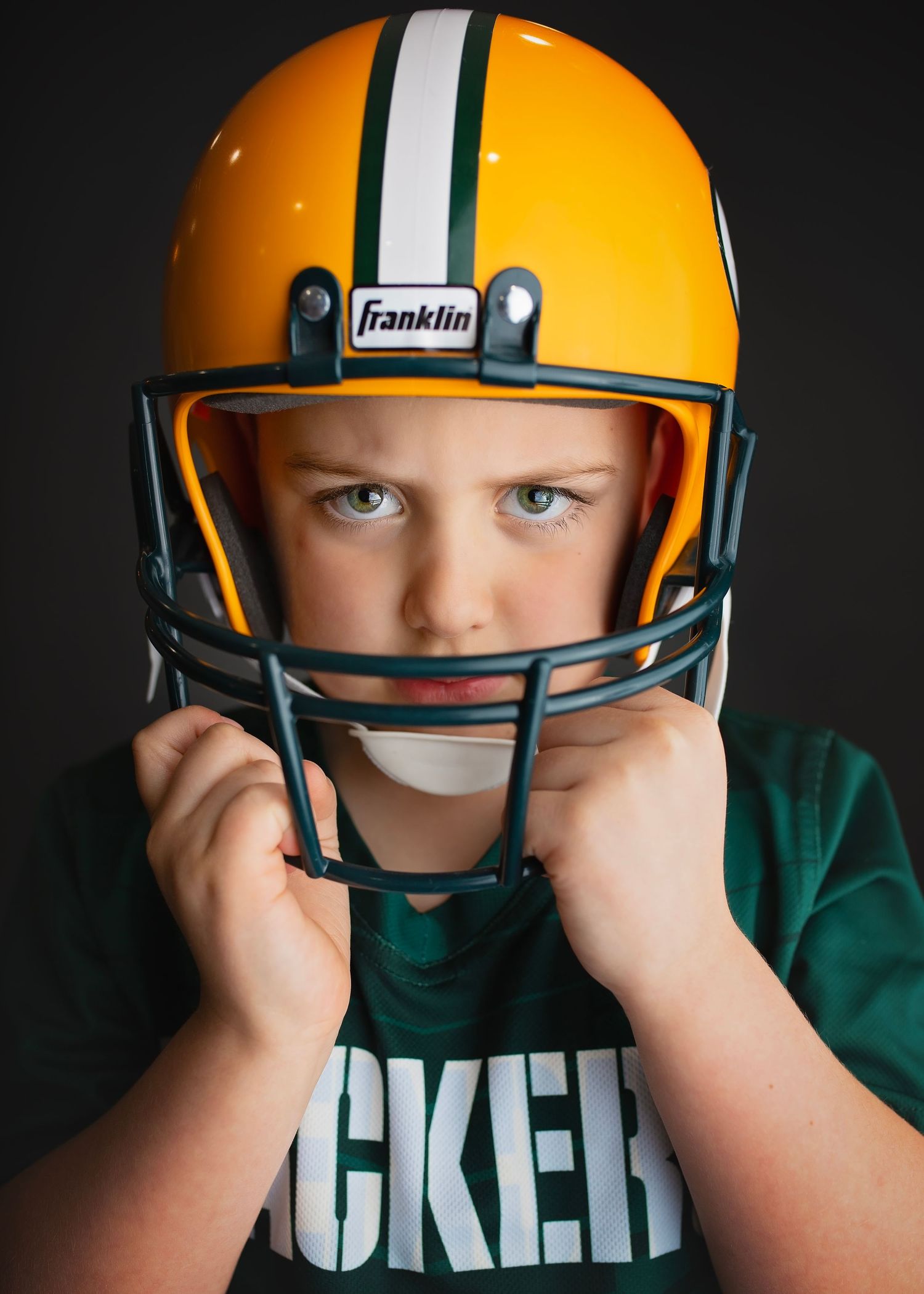Young boy wearing Green Bay Packers Franklin football helmet in intense pose on dark background.