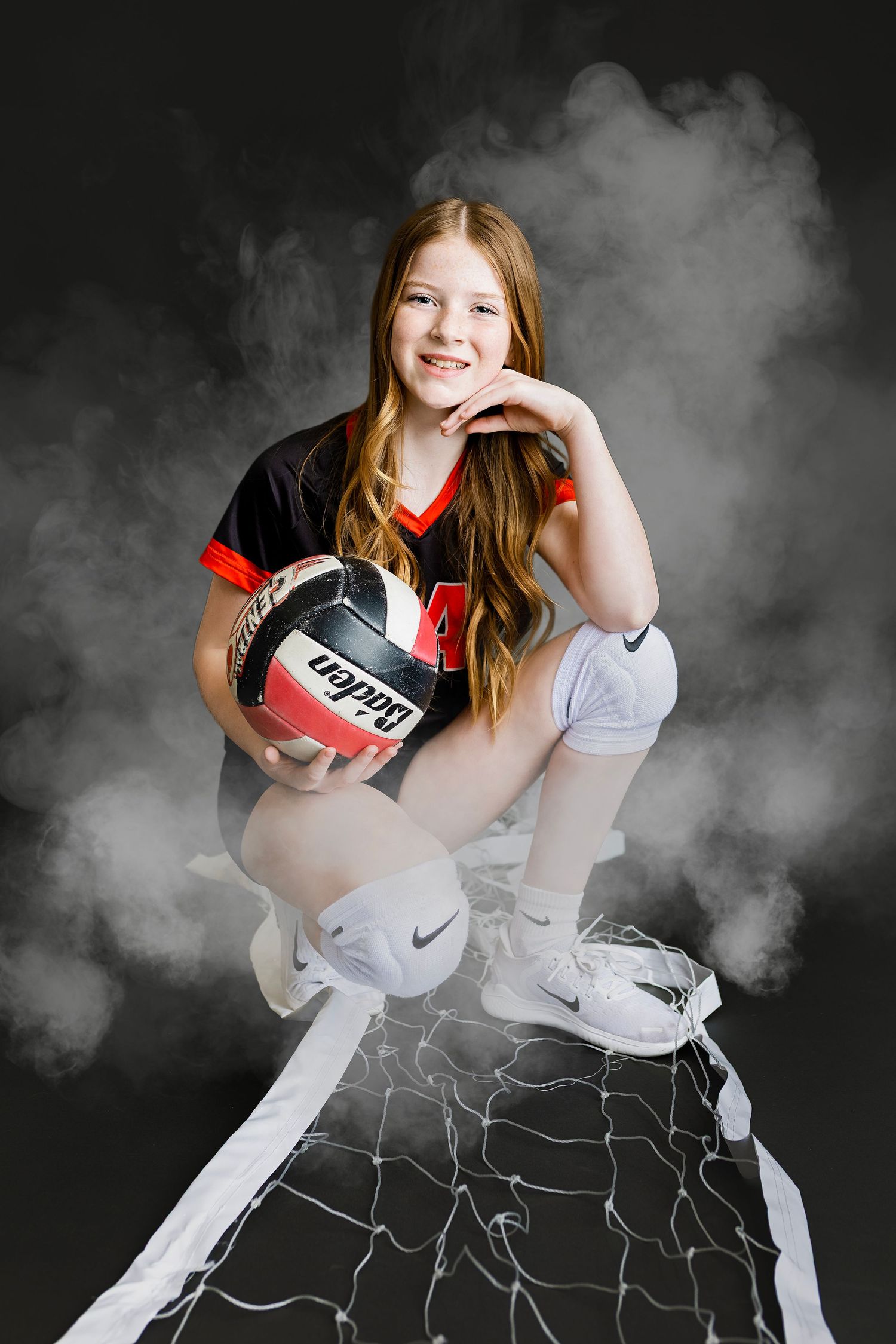 Young redheaded girl in volleyball uniform crouches on a net holding a Baden ball with dramatic smoke background.