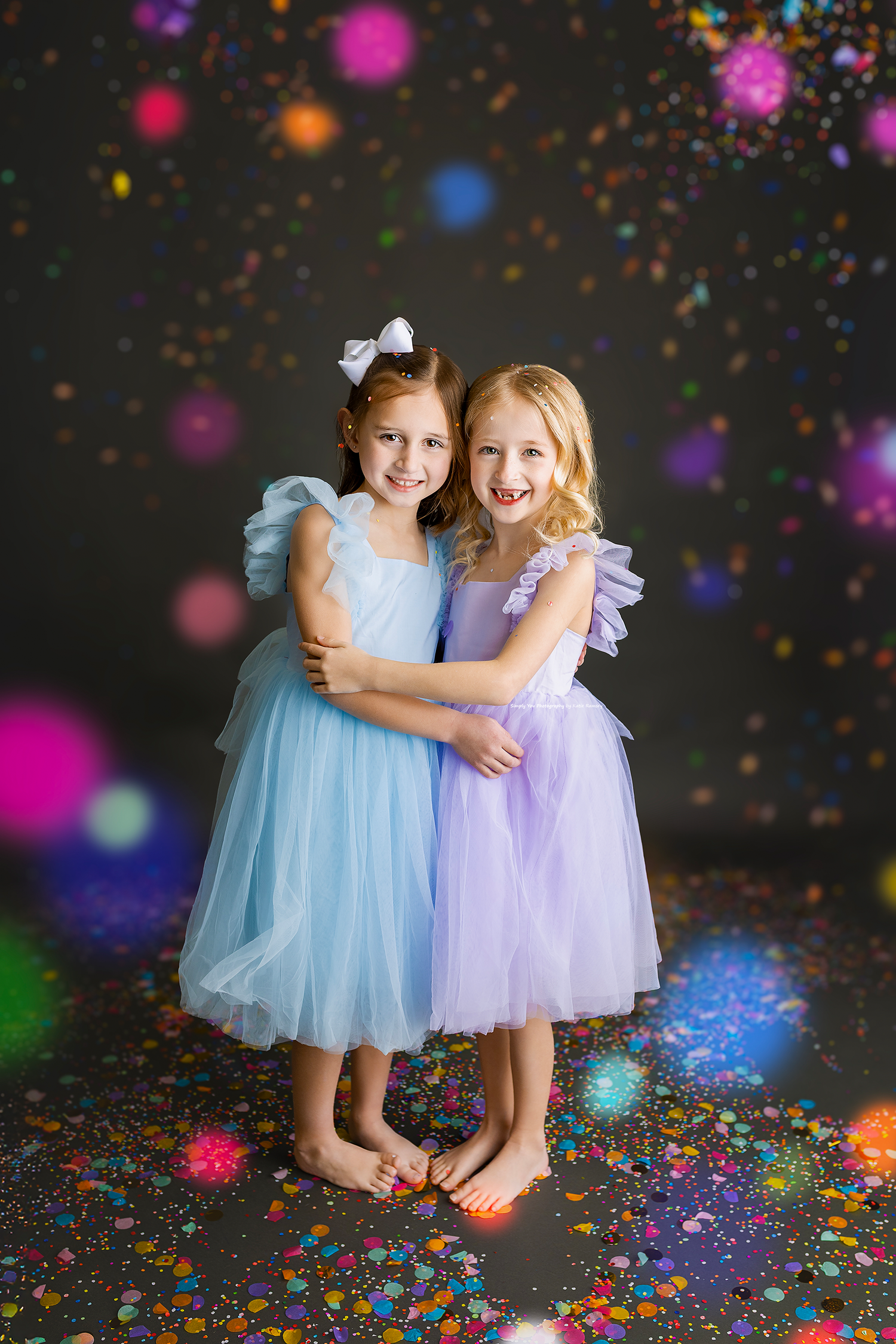 Two girls in blue and purple tulle dresses hugging surrounded by colorful confetti.