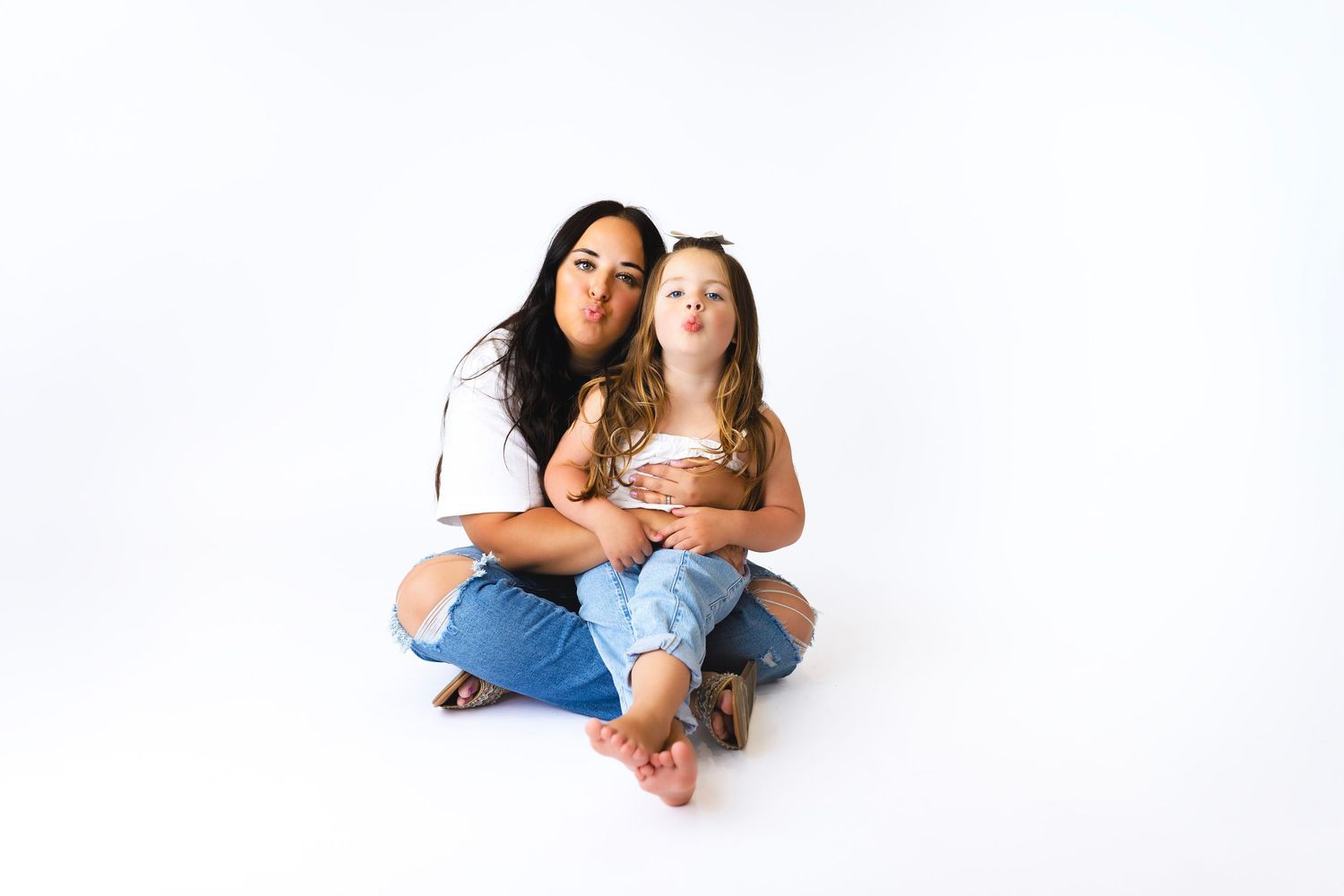 A woman and young girl sit cross-legged on white backdrop, both making silly faces in matching white tees and jeans.
