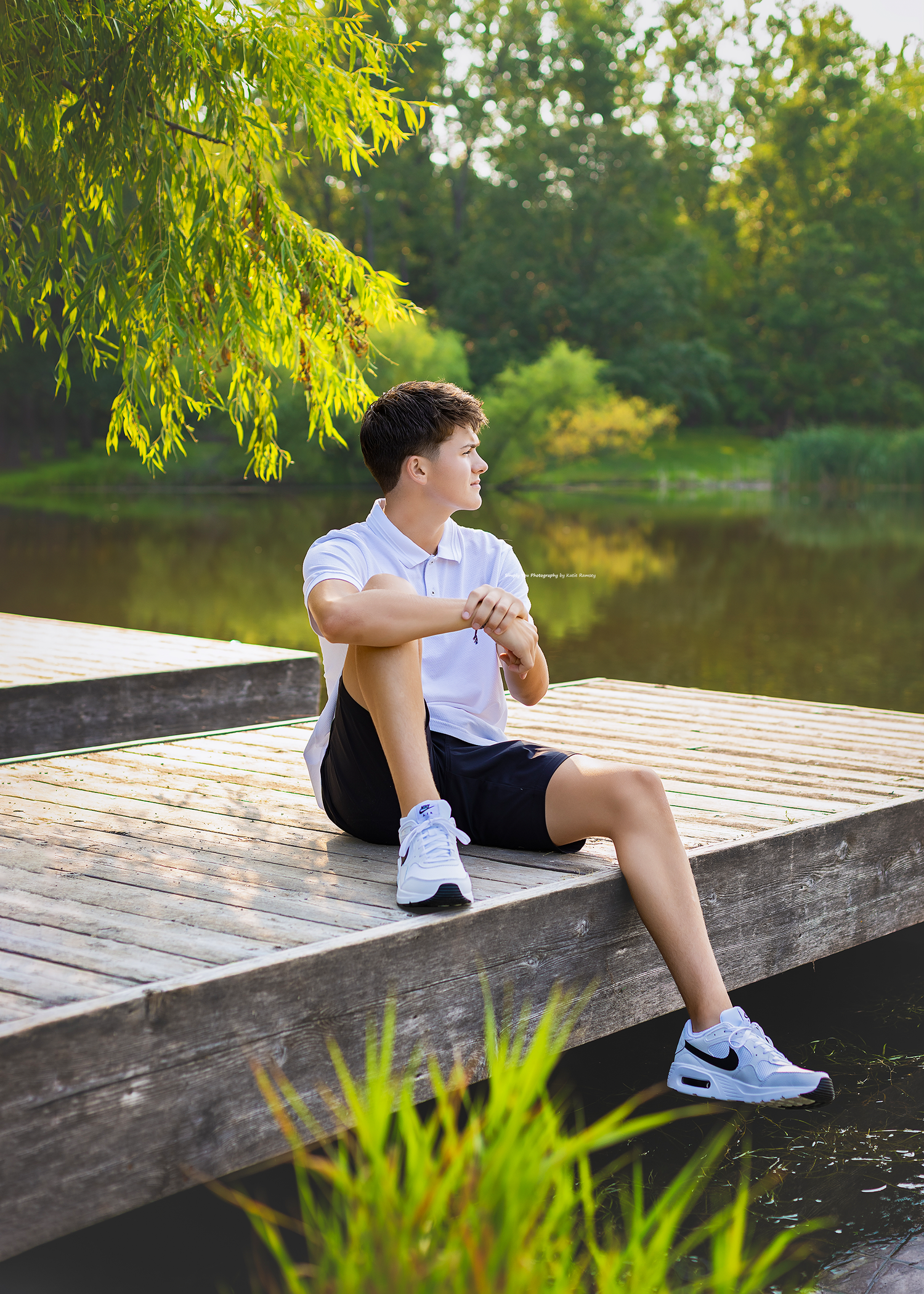 Young man in white polo and black shorts sitting on a wooden dock by a serene lake surrounded by lush green trees.