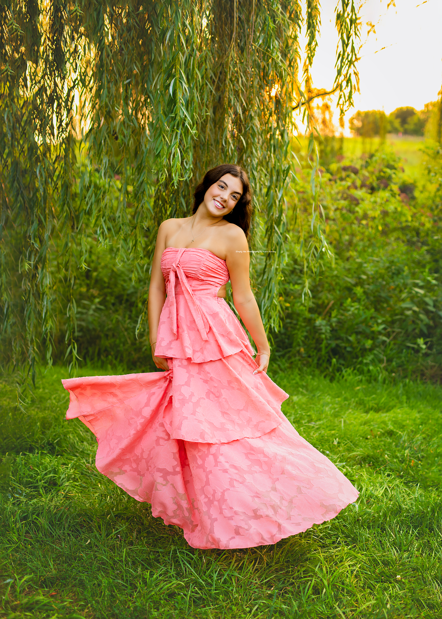 A young woman twirls in a flowing pink strapless gown outdoors under a weeping willow tree at golden hour.