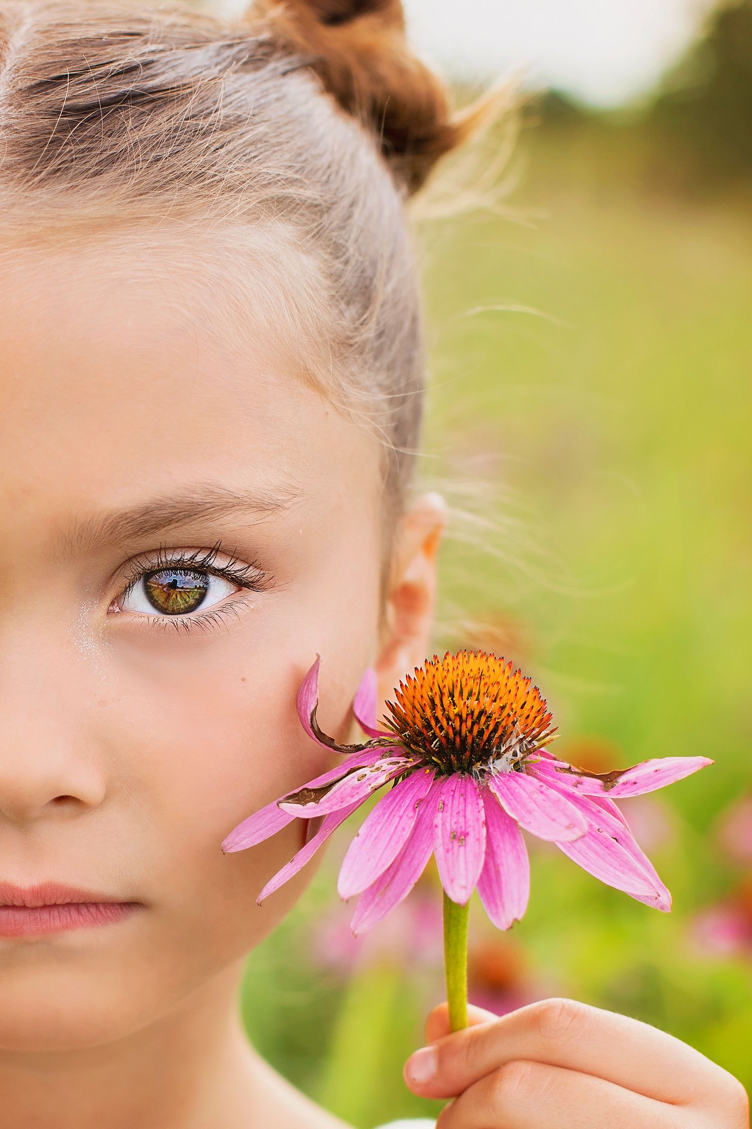 Close-up of a young girl holding a pink echinacea flower near her face in a garden.