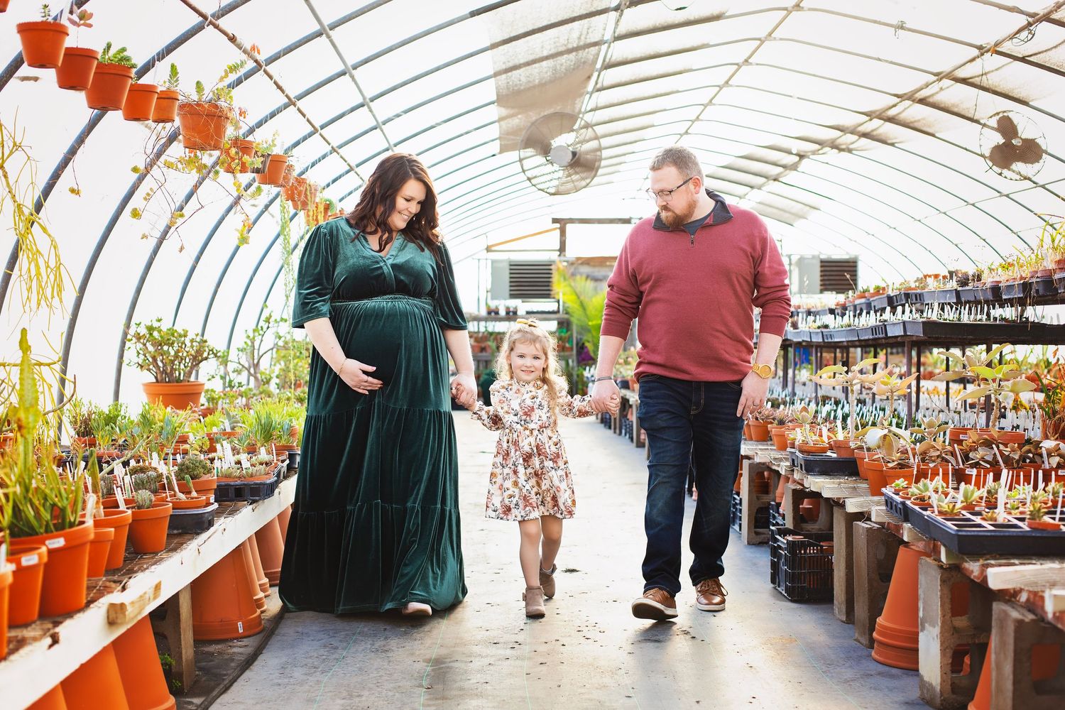 Pregnant woman in green velvet dress walks hand-in-hand with toddler and husband through plant greenhouse.